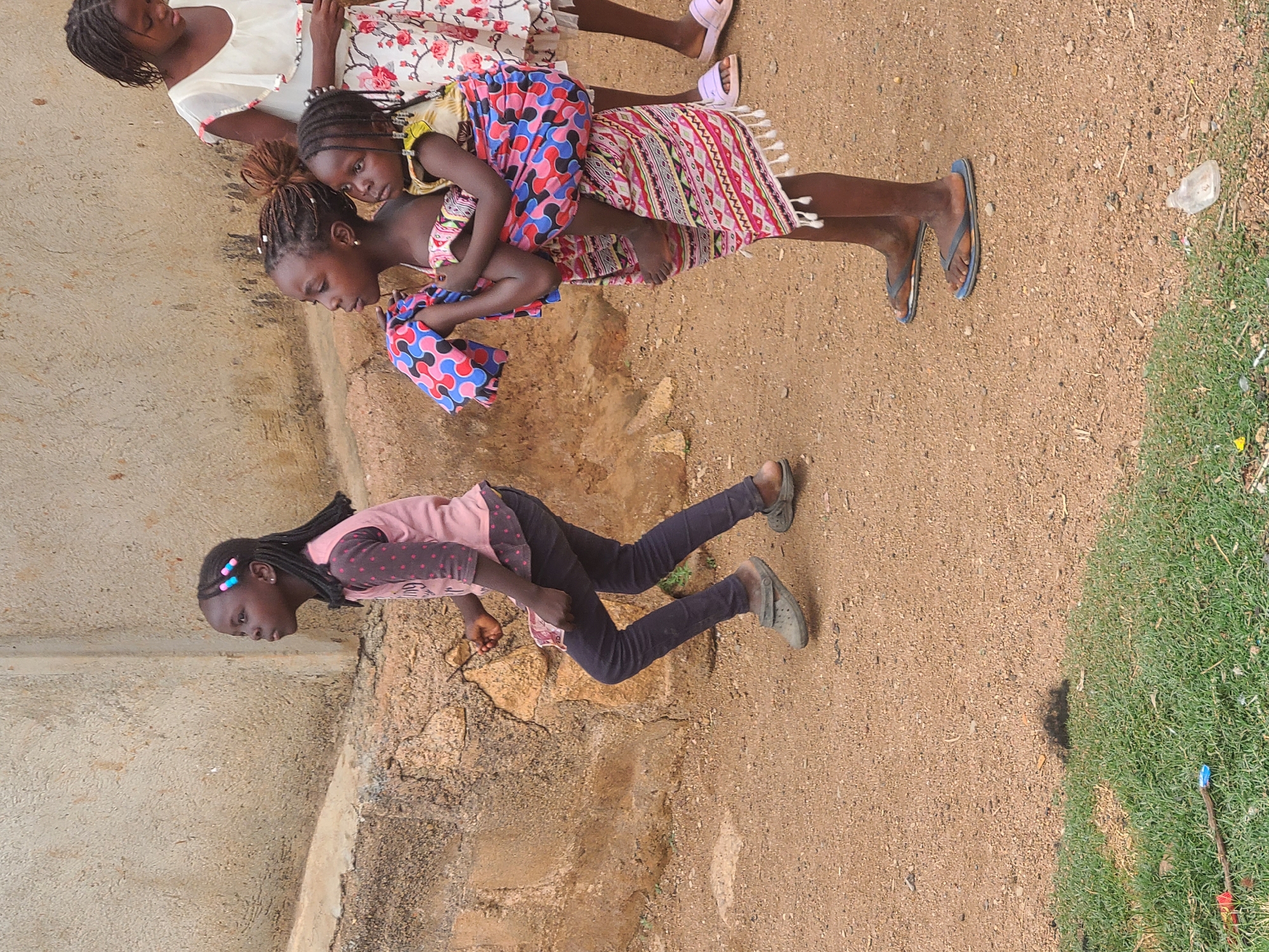 The image depicts an outdoor scene in Jos, Nigeria. Three young girls are centrally located. One girl is carrying a younger girl on her back, while another girl stands separately. The children are of African descent and appear to be playing. They are dressed in casual clothing; the girl carrying the other wears flip-flops. Another child, partially visible, is wearing a white dress with a floral pattern. The standing girl wears dark pants, a pink shirt, and gray shoes.
The environment consists of a dirt ground with some patches of green grass on the right side. In the background, there is a weathered concrete wall. There appears to be some erosion of the ground with exposed soil and rocks beside the wall. Debris, including a discarded plastic bottle, is visible in the background.
