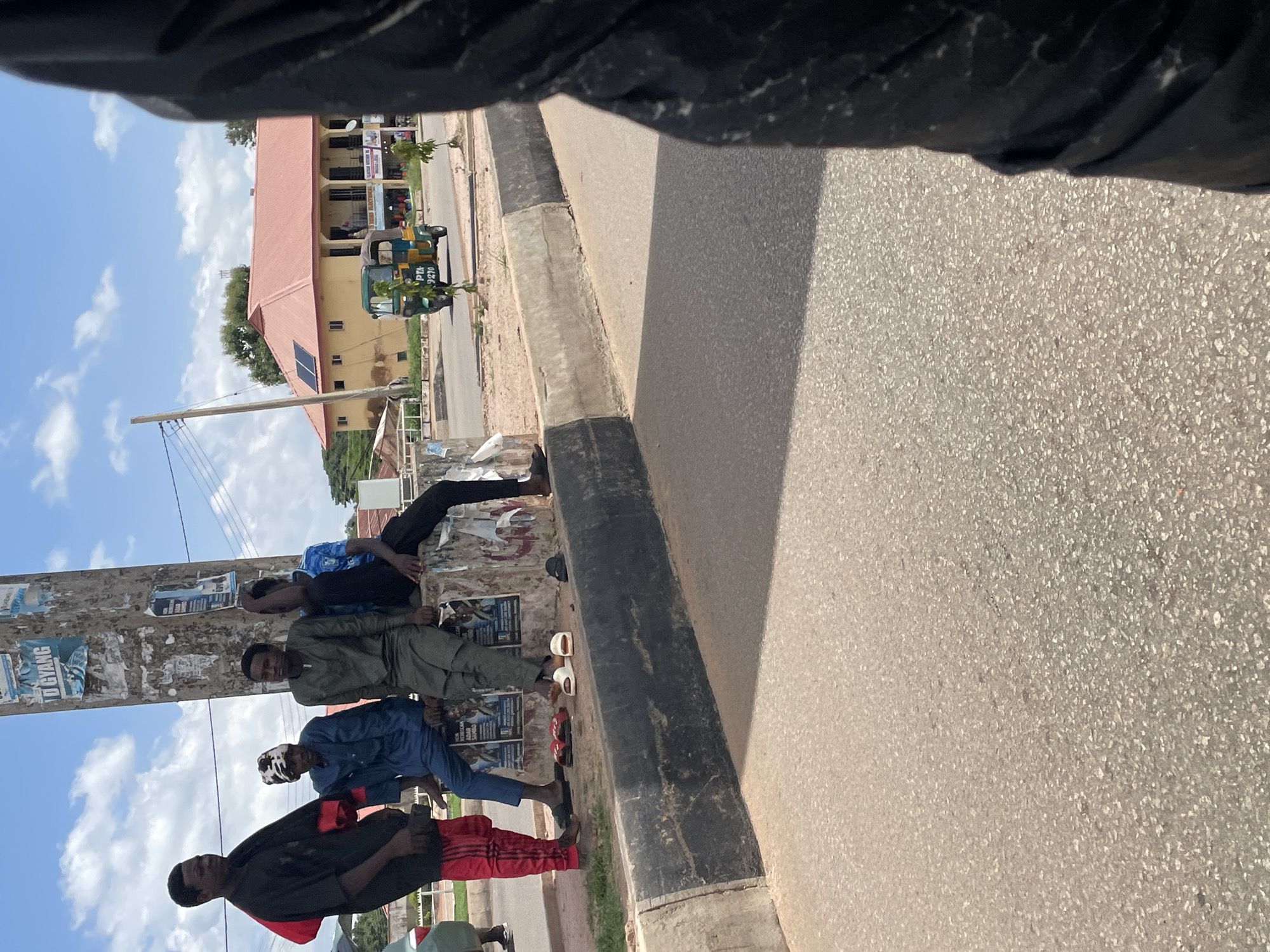 A group of young men are gathered on a street in Gura Top, Nigeria, under a bright, cloudy sky, suggesting it is daytime. The scene appears to be a roadside, with a paved road and a sidewalk. Some of the men are sitting or standing near a utility pole covered in posters. Their clothing is casual, with some wearing traditional attire and others more modern styles.
One man in the foreground wears a black shirt with red accents and red and black striped pants. Another in a blue outfit sits near him. Further down, a man in a green garment is seated, and another in a blue shirt is seen with his head down. A few individuals are standing, observing their surroundings or interacting with each other. White and red sandals are visible on the ground.
In the background, a building with a red roof and yellow walls is visible, along with trees and a small, three-wheeled vehicle, possibly an auto-rickshaw. The overall atmosphere seems relaxed, with no overt signs of distress or urgency. The presence of posters on the pole suggests a public area where announcements or advertisements might be displayed. The light and shadows indicate a sunny day. The scene captures a moment of everyday life in an urban or semi-urban setting.