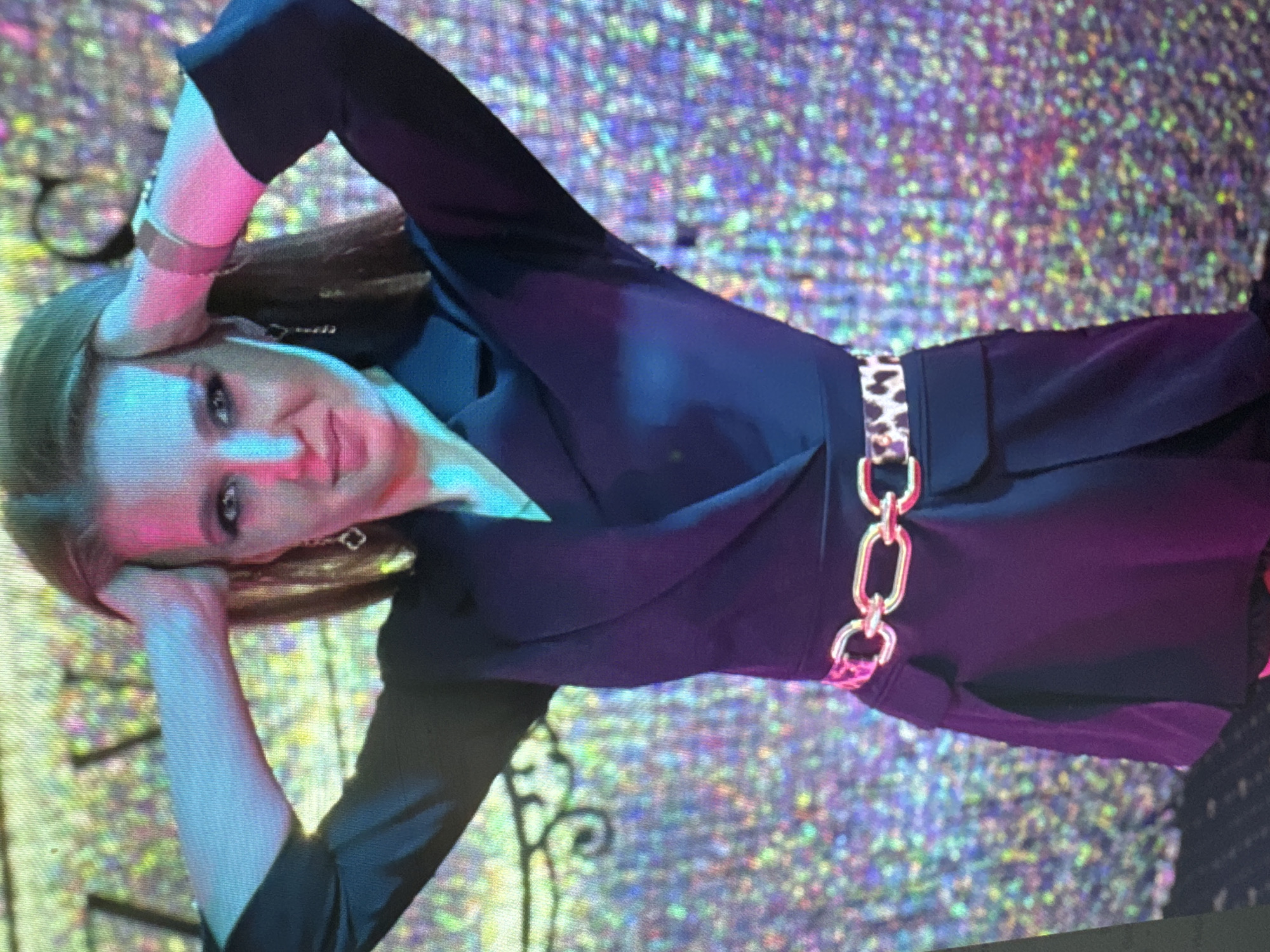 A young woman with long, brown hair is standing in front of a sparkly, iridescent background. She is wearing a black blazer dress with a leopard print belt and a gold chain belt. Her arms are raised, and her hands are resting on her head. She is looking at the camera with a slight smile. The lighting is a mix of blue and pink.