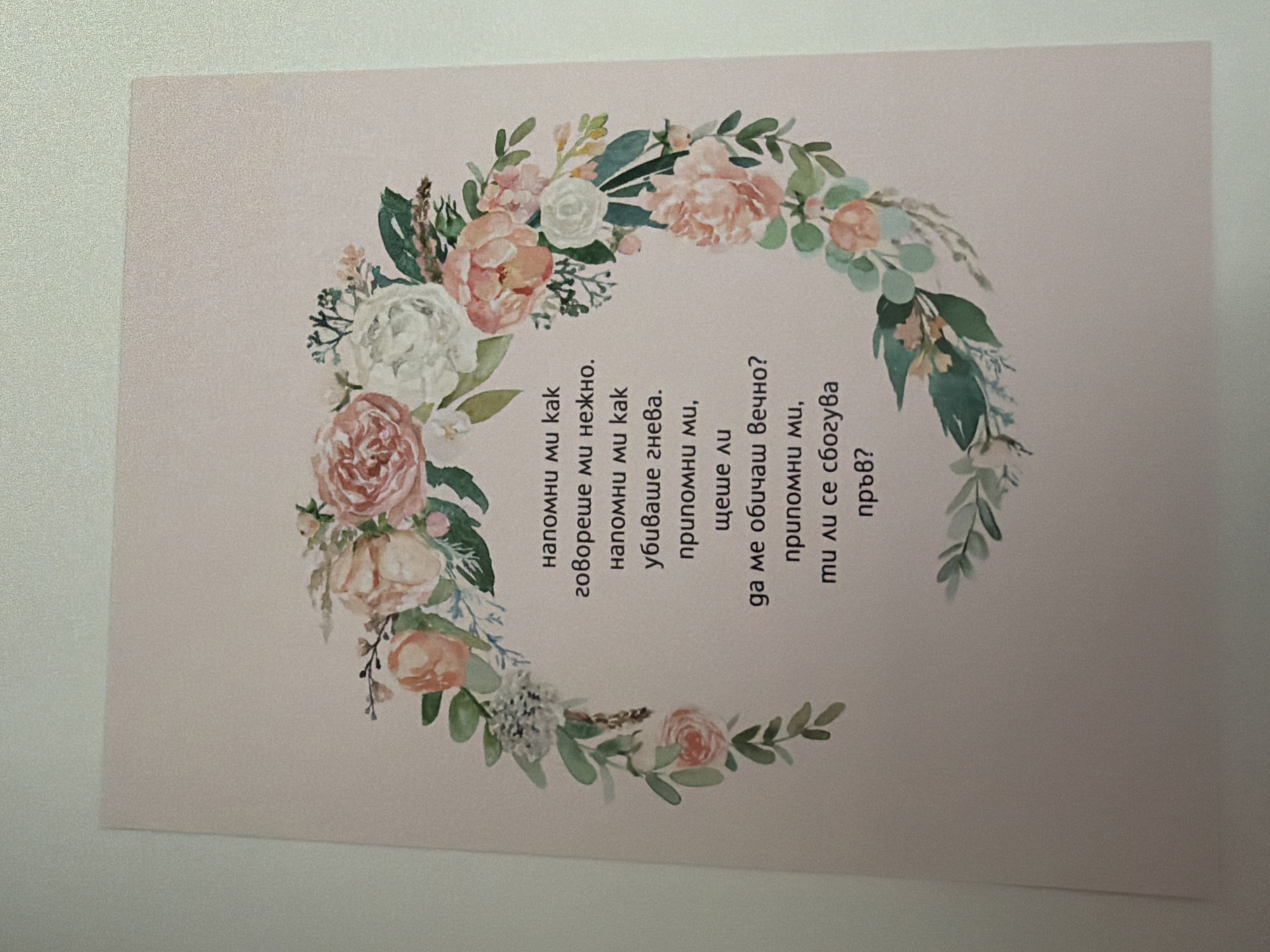 The image shows a postcard with a floral wreath design surrounding text in Bulgarian. The text appears to be a poem or a message. The postcard is a light pink color and is placed on a white surface, possibly a table or desk. The lighting suggests it was taken indoors. There are no people visible in the image. The floral wreath consists of various flowers, including roses in shades of pink and white, and greenery. The text is centered within the wreath. There is no identifiable location within the city of Pazardzhik, Bulgaria, as the image is a close-up of the postcard itself and its immediate surroundings, which are not specific to any particular place.