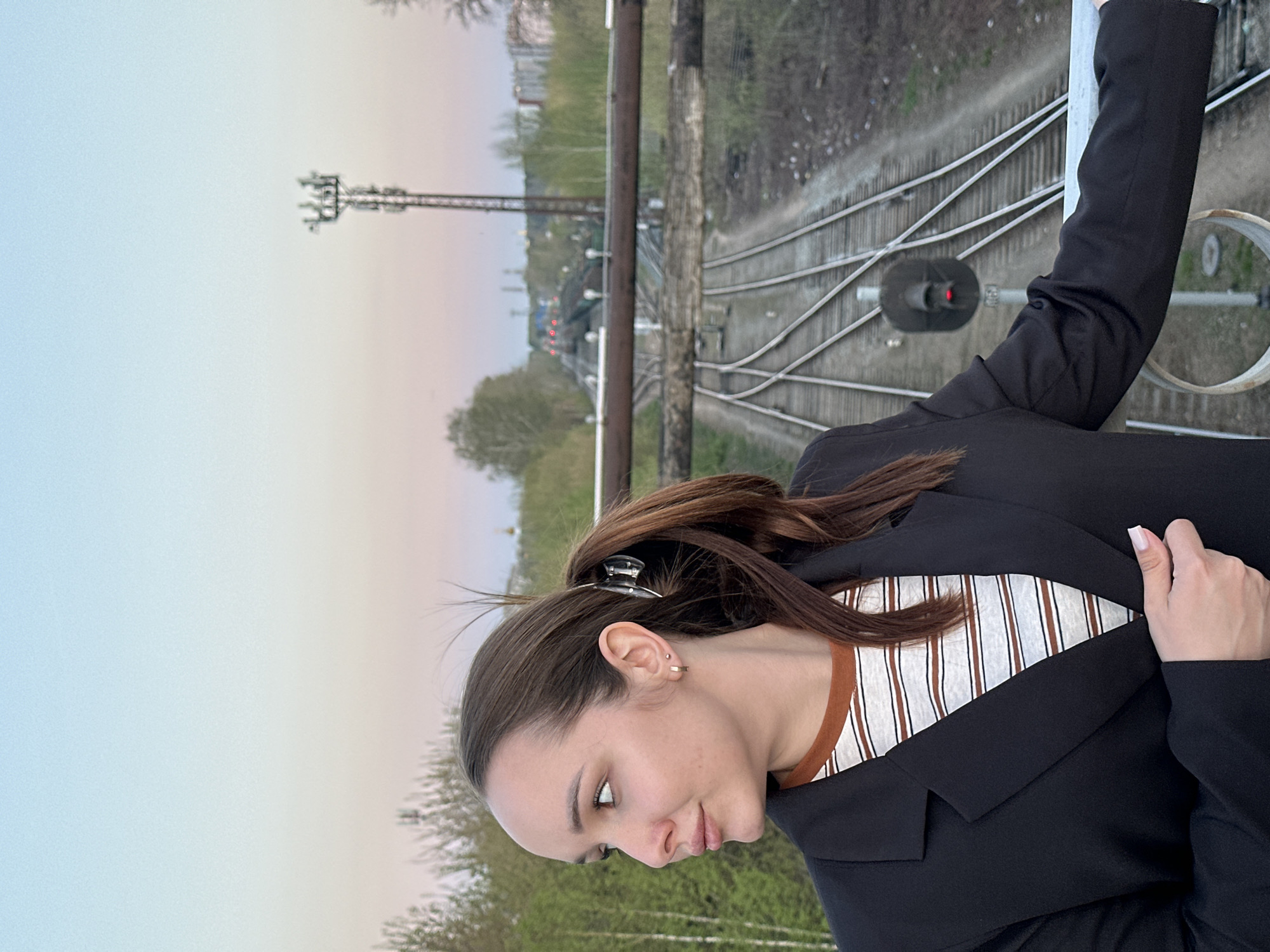 A medium shot, taken from above, depicts a woman standing on a bridge or overpass, with railway tracks visible below. The woman, wearing a black blazer over a striped shirt, is holding onto a railing of the structure. Her hair is pulled back with a clip, and she has an earring in her left ear.
The background shows a complex of railway tracks, signals, and overhead lines. In the distance, trees and buildings indicate an urban environment. The sky transitions from light blue to a soft pink hue near the horizon.
Based on the visible infrastructure and the overall composition of the scene, the location is plausibly the railway tracks area of Chaikovsky, Russia, although specific landmarks for precise confirmation are absent.