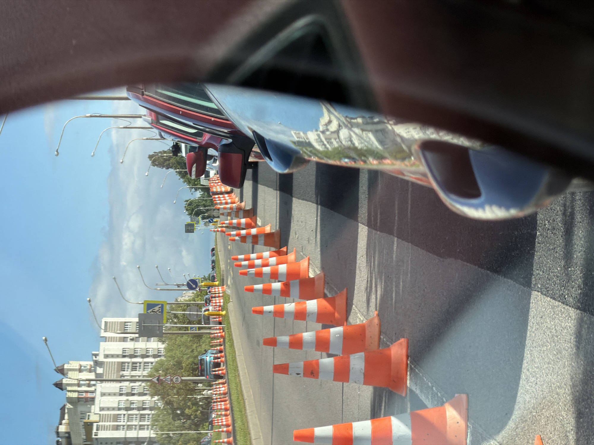 An outdoor, eye-level shot captures a road with numerous orange and white striped traffic cones, indicating a closed lane or construction. The cones line up along the right side of the road, and also form a divider between the current lane and what appears to be a closed lane. In the background, several cars are visible, some parked and some in motion. Beyond the road and cars, a large, multi-story building with a light-colored facade and a distinctive rooftop structure stands tall against a bright, partly cloudy sky. Streetlights are spaced along the roadside. The image is taken from inside a vehicle, with the side mirror of a red car and a portion of the car's interior visible in the foreground, blurring the view of the scene. The overall impression is one of urban infrastructure and traffic management. This scene is likely from Minsk, Belarus, given the architectural style of the buildings and the common use of such traffic cones for road work or events.