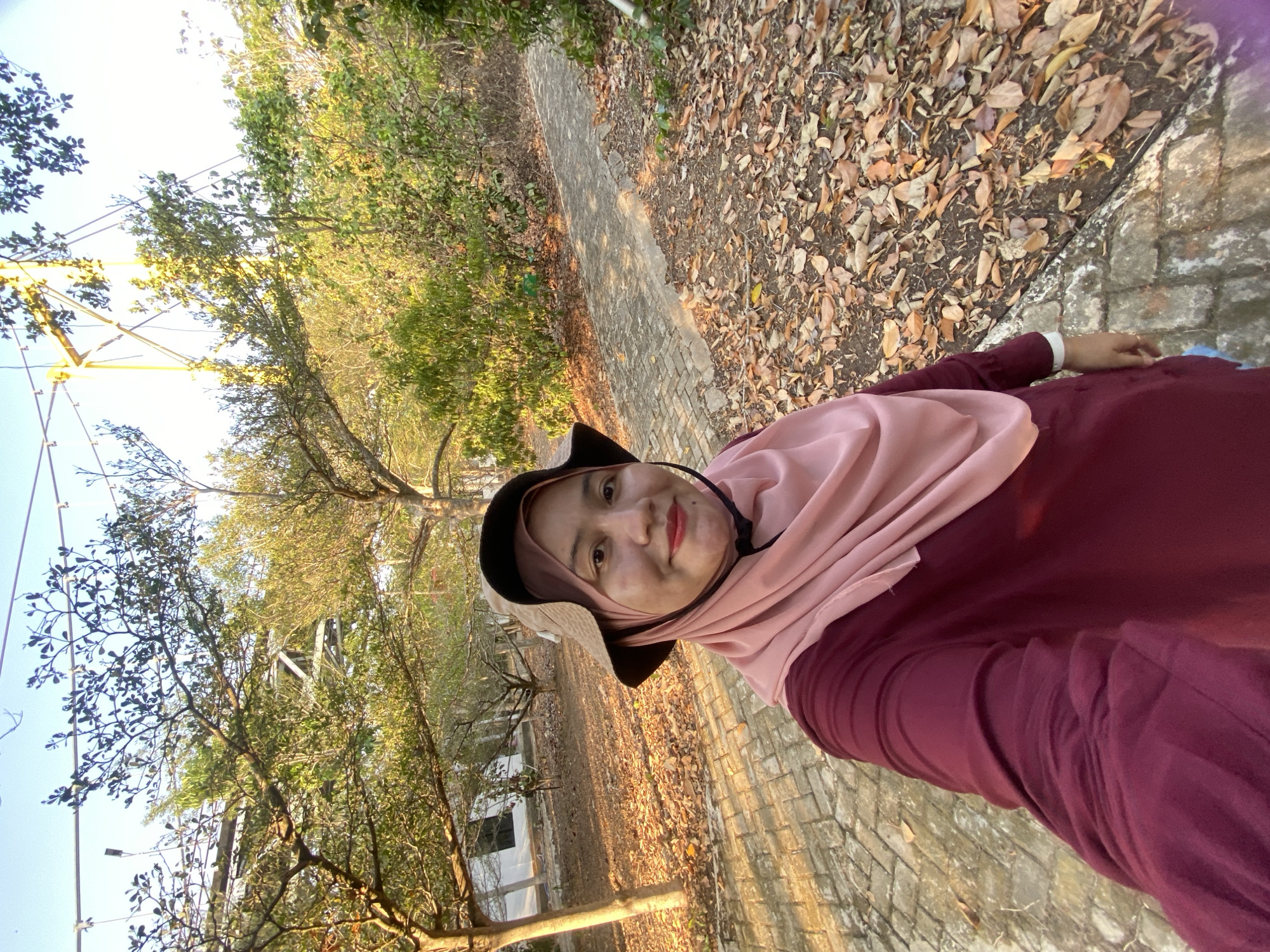 A medium-shot, outdoor, low-angle selfie shows a woman standing on a paved walkway with fallen leaves. The woman, wearing a maroon long-sleeved shirt, a light pink hijab, and a wide-brimmed brown hat, smiles at the camera. The walkway is constructed of light-colored rectangular bricks. Trees with green and yellow foliage line the path, and some bare branches are visible. In the background, a white building with dark windows is partially obscured by trees. Overhead, metal support structures for what appears to be a bridge or zipline are visible against a clear blue sky. The lighting suggests it is late afternoon, with warm sunlight casting shadows. The overall environment appears to be a park or recreational area.