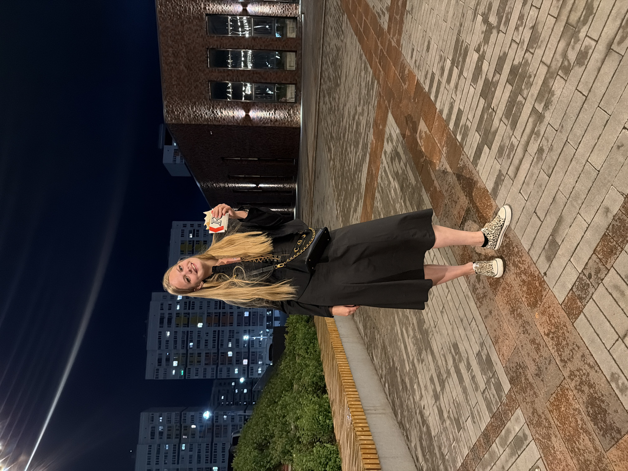 A young woman with blonde hair stands in an urban plaza at night, holding a wrapped food item. She is wearing a black jacket, a black skirt, and leopard-print sneakers. The plaza features a brick-patterned walkway, a wooden bench, and lush green bushes. In the background, several apartment buildings with illuminated windows are visible, along with a modern brick building. The scene is characteristic of Pervomaysky, Russia, where such mixed residential and modern architectural styles are common. The lighting suggests it is evening or nighttime.