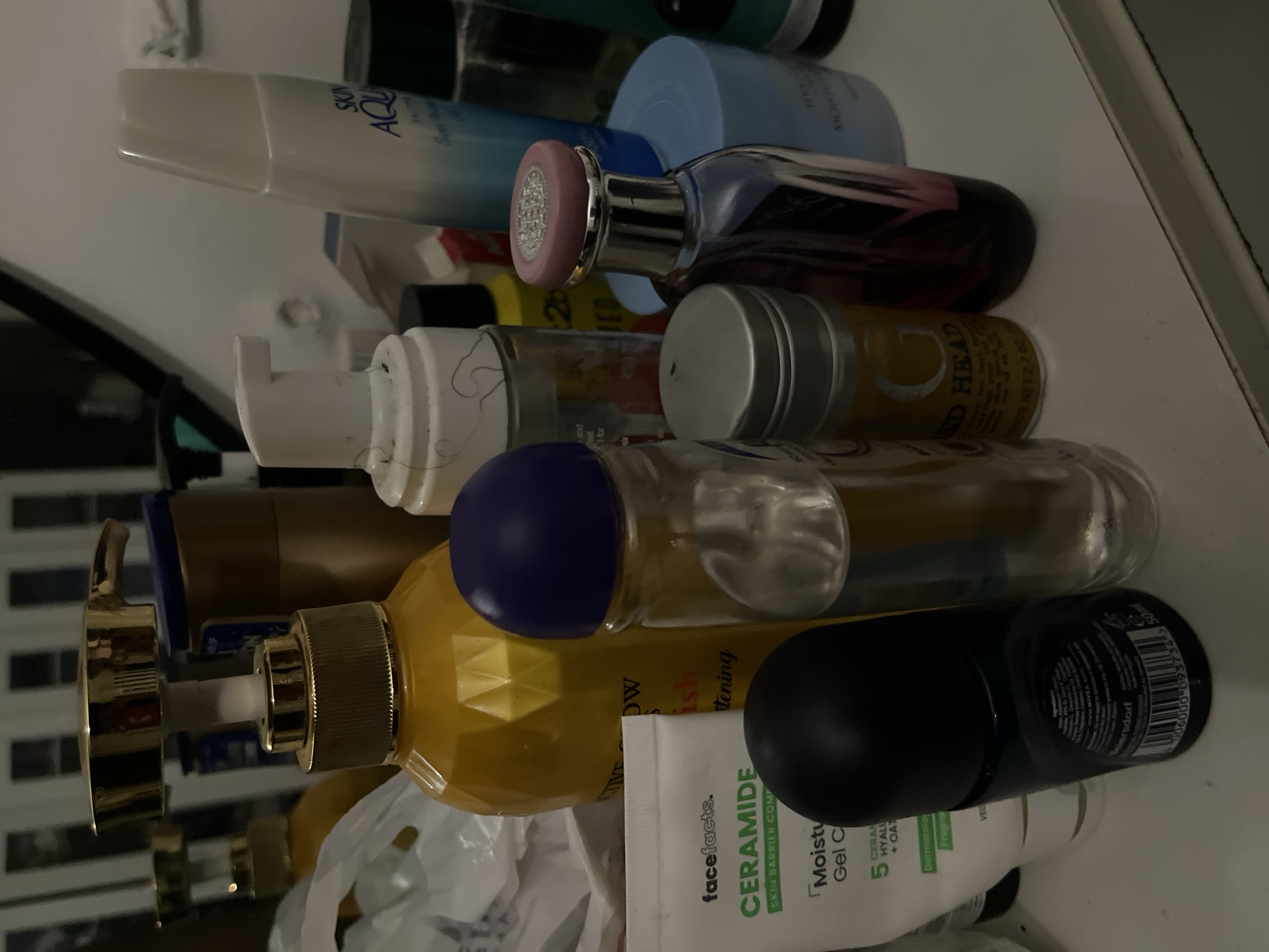 The image shows a collection of various cosmetic and skincare products arranged on a white surface, likely a shelf or countertop. The items include bottles, pumps, and jars, suggesting a personal grooming or beauty collection. The lighting is somewhat dim, creating shadows and highlighting the textures of the products. The background is blurred, but appears to be an indoor setting with vertical elements that could be shelves or furniture. There are no people or actions depicted in the image. The specific location in Abuja, Nigeria, cannot be determined from the visual information provided in the image.