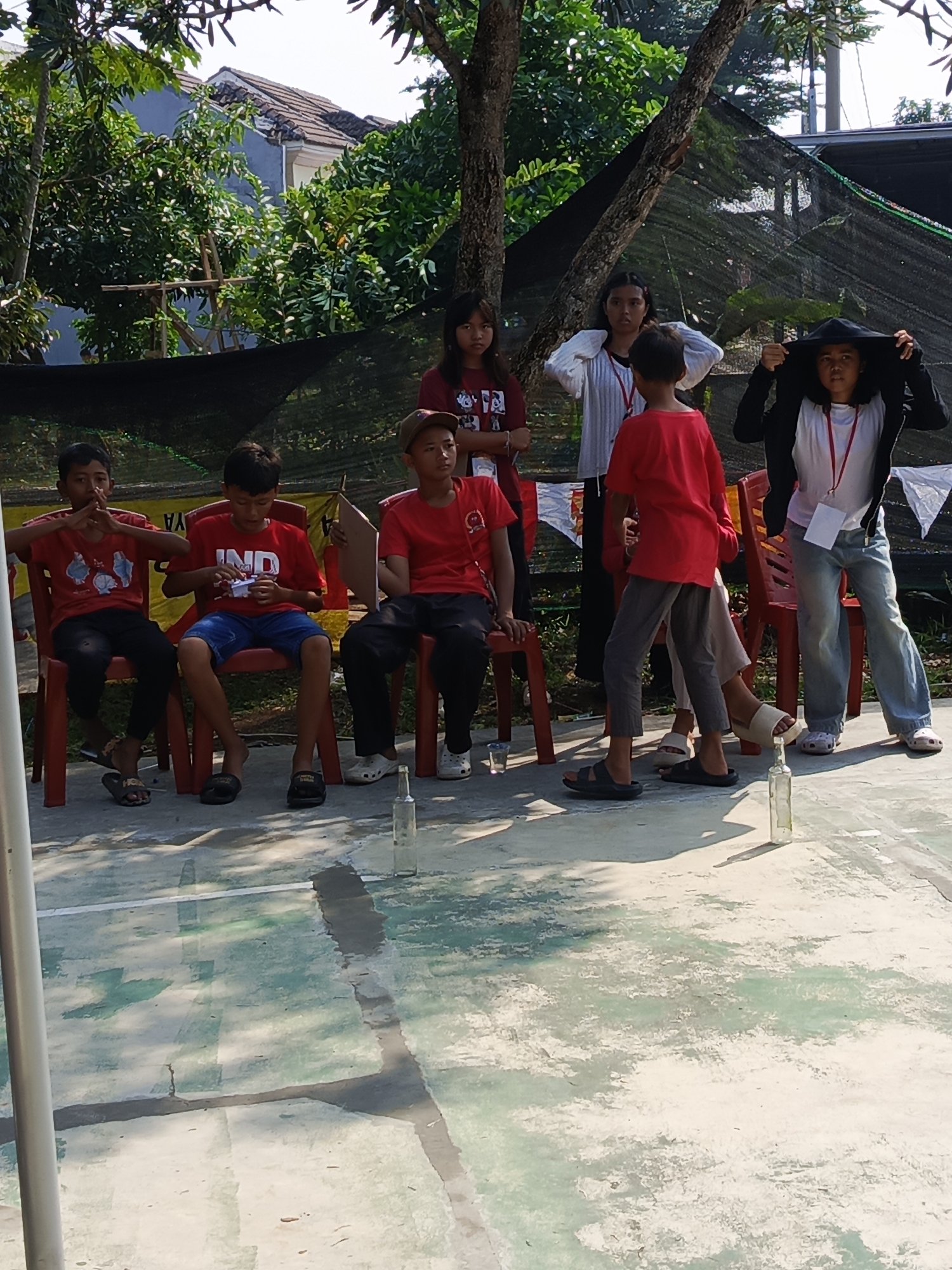 The image depicts a group of young people, predominantly children and teenagers, gathered outdoors in what appears to be a recreational or community setting in Ciseeng, Indonesia. The foreground shows a concrete ground with white lines, possibly a court or paved area. Several clear glass bottles are placed upright on the ground. In the middle ground, a line of three boys and one girl are seated on red plastic chairs. The boy on the far left is wearing a red t-shirt and black pants. The second boy from the left, also in a red t-shirt and blue shorts, is looking down at his hands. The third boy from the left is wearing a red t-shirt and dark pants, holding a piece of paper or a card. The girl, on the far right of this seated group, is wearing a white t-shirt and jeans. Behind them, standing and in motion, are four more individuals. Two girls are standing together; one in a white long-sleeved shirt and the other in a red t-shirt. A boy in a red t-shirt is walking towards the viewer, and another person with a hood pulled up is standing near the edge of the frame. A black mesh screen or net is visible in the background, with trees and foliage also present. The overall impression is of an outdoor activity or event possibly involving a game.