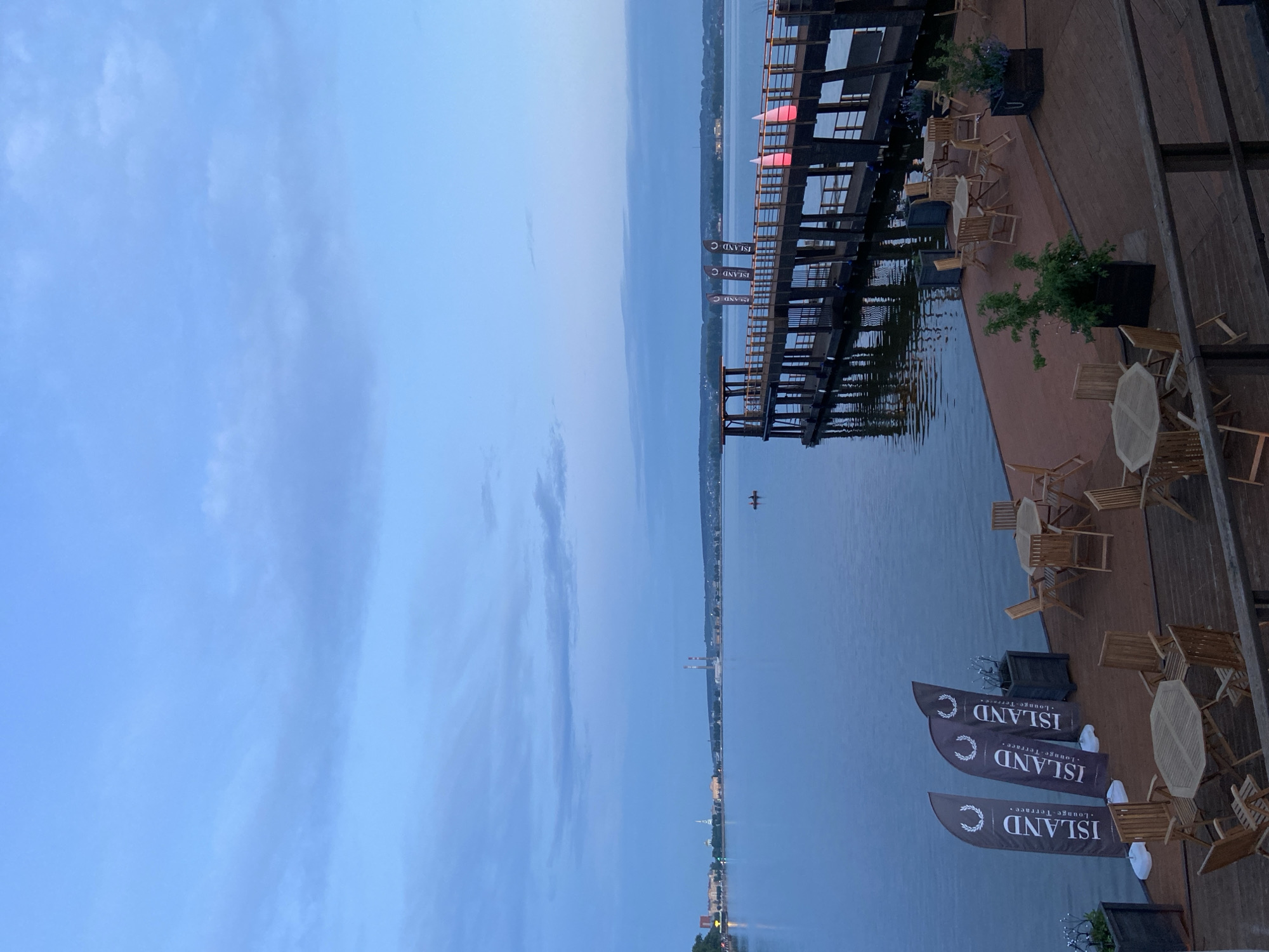 This is a wide-angle, eye-level, outdoor, daytime photograph of a waterfront area in Votkinsk, Russia, captured at dusk. The scene features a large body of water, likely the Votkinsk Reservoir, under a cloudy, pale blue sky.
In the foreground, a wooden deck area is laid out with numerous tables and chairs, made of light-colored wood. Some tables are octagonal, others round, and all are surrounded by folding chairs. Three tall, feather flags with the logo "ISLAND Lounge Terrace" are positioned on the left side of the deck. Planters with greenery are also visible.
A dark wooden pier extends from the right side of the frame into the water. The pier has a covered structure at its end and is lined with railings and decorative red lights. A small red boat with two occupants can be seen on the water in the distance, in front of the pier.
Across the water, the Votkinsk skyline is visible. It includes buildings, structures, and what appear to be industrial chimneys with red-and-white striped tops. The water's surface is calm, reflecting the muted colors of the sky and structures. The overall atmosphere is tranquil, suggesting the beginning of an evening at an outdoor venue.