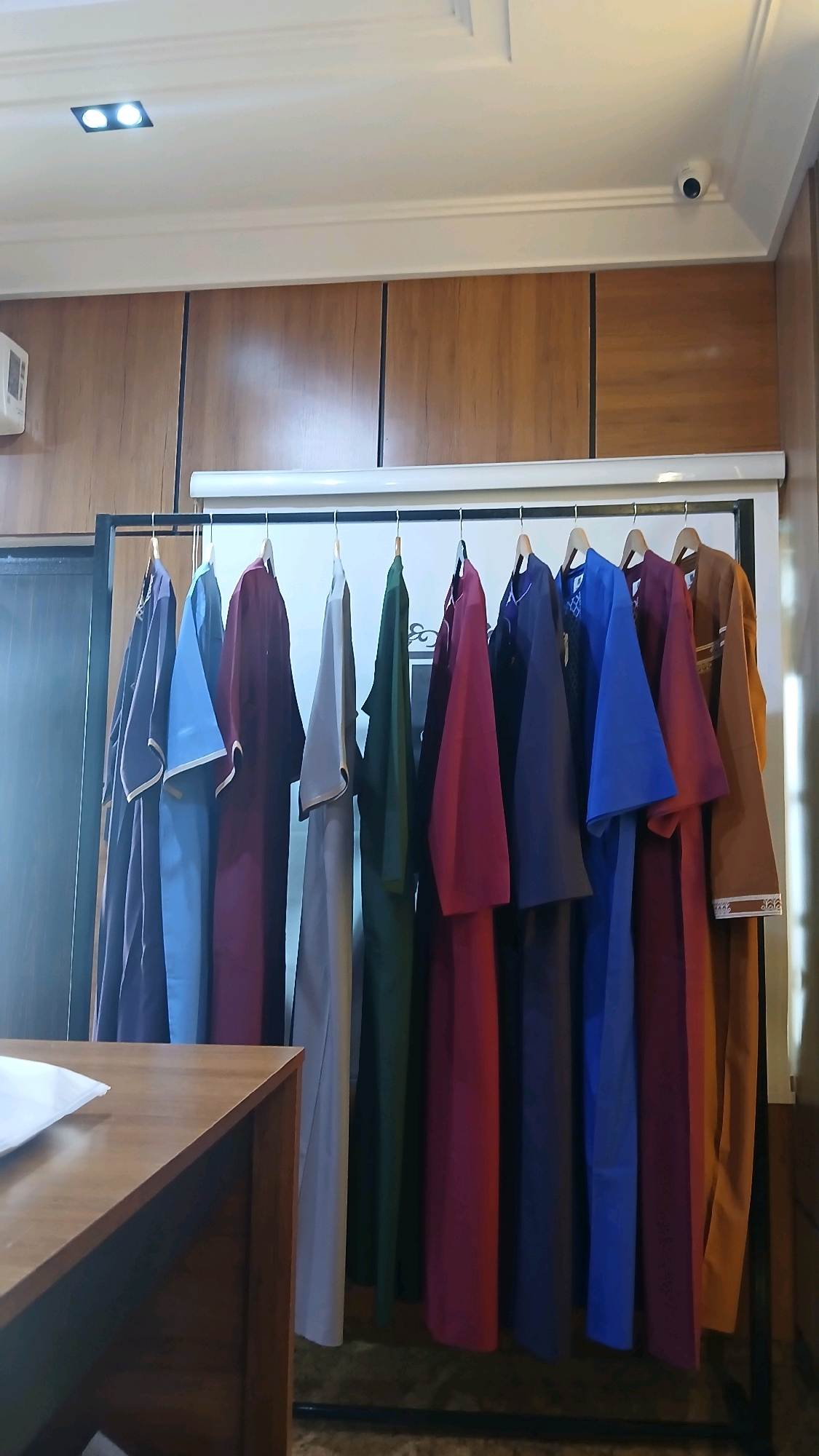 A rack of long, flowing garments in various colors hangs against a wood-paneled wall in what appears to be a retail setting or storage area. The garments include shades of purple, blue, maroon, gray, green, and a light brown or tan. They are hung on wooden hangers. Above the rack, a white shelf or ledge is visible, and further up, the white ceiling features recessed lighting and a white security camera. A portion of a wooden table or counter is in the foreground on the left. The overall impression is one of organized inventory, likely clothing. There is no discernible indication of the specific location being Barikin Jirga Summa, Nigeria.
