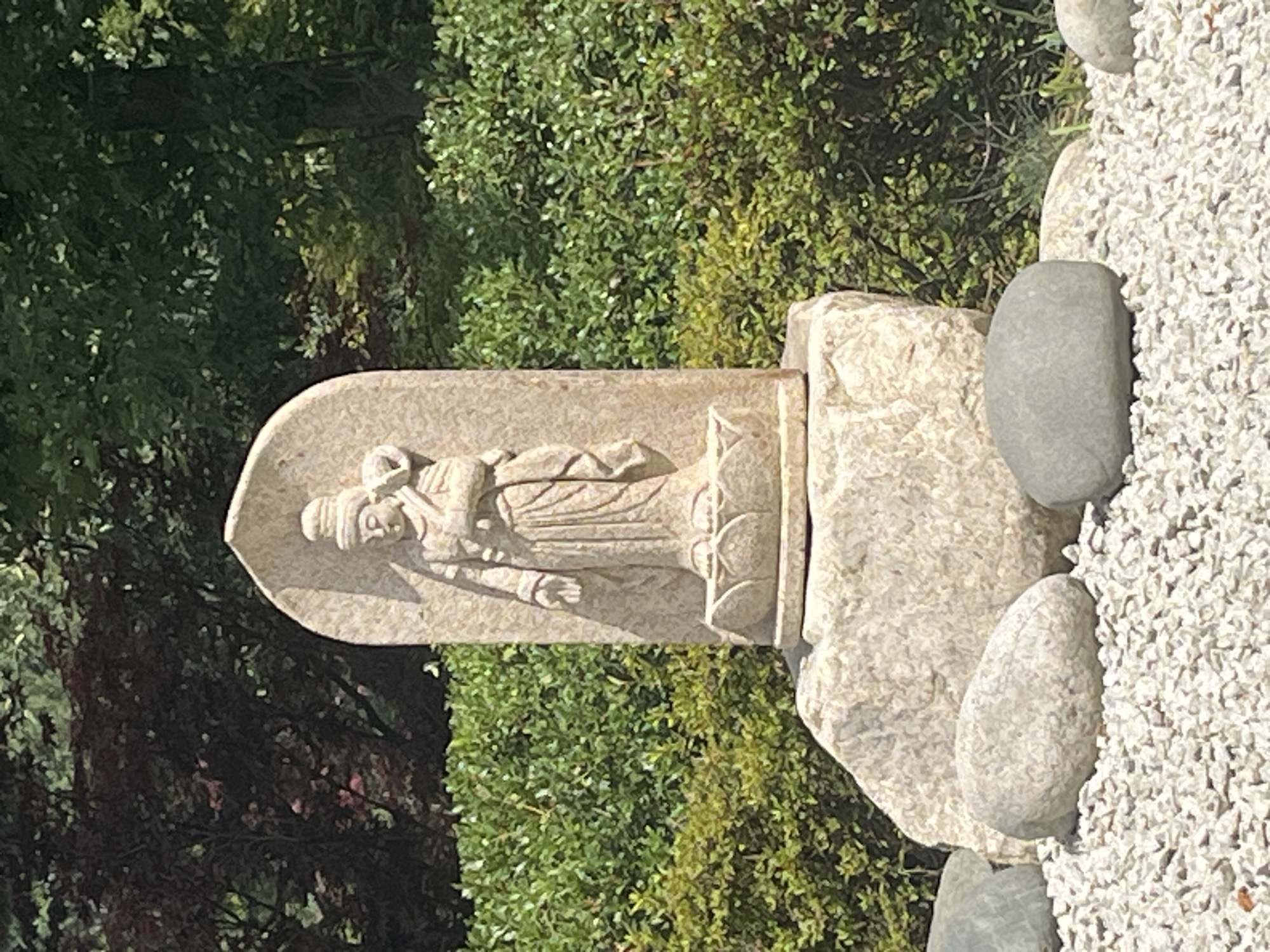 A stone statue of a Buddhist deity, possibly Kannon, stands on a pedestal atop a large rock. The statue is carved from light-colored stone and depicts the deity in a flowing robe, holding an object in its left arm. The pedestal features lotus petal detailing. The statue is surrounded by lush green bushes and trees, with a background of darker foliage. In the foreground, white gravel and several smooth, grey stones are visible. The scene appears to be outdoors, likely in a garden or park setting. There is no identifiable information about the specific location in Krasnodar, Russia.