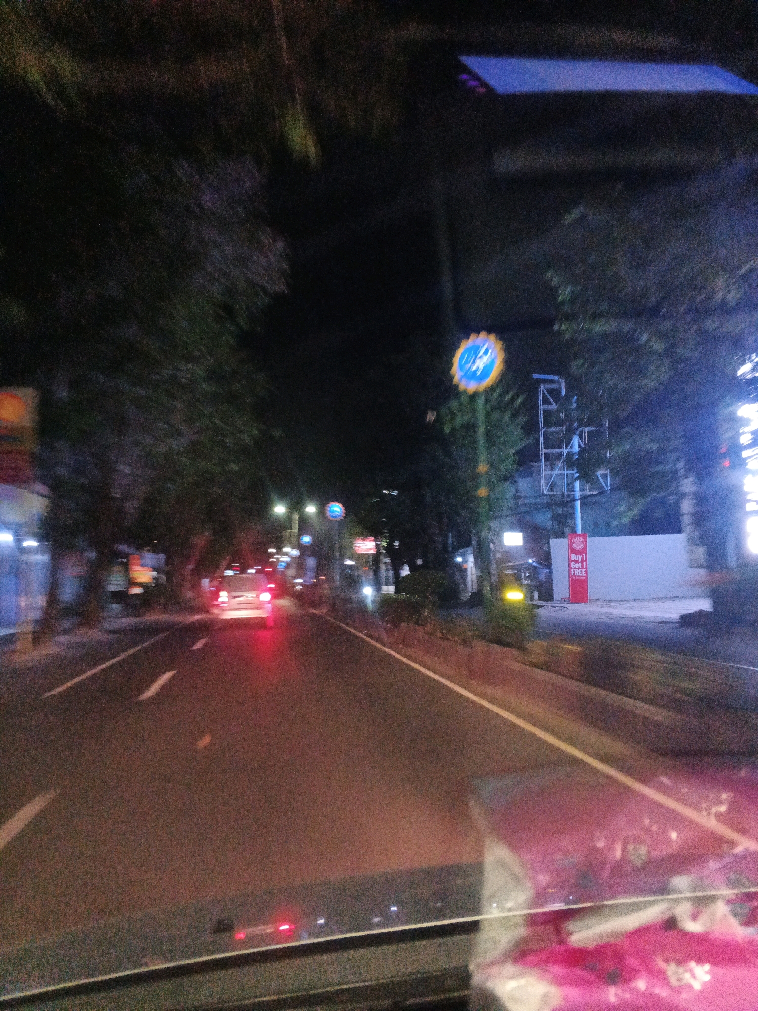 The image captures a nighttime view from a moving vehicle, likely a car, on a city street in Jatisari, Indonesia. The road is paved with clear lane markings. On the right side of the road, there are trees lining the street. A blue and yellow sign with a bicycle symbol is visible on a pole. Further down the street, a tall structure resembling a utility pole or scaffolding is present, with a red sign that says "Buy 1 Get 1 FREE". The headlights and taillights of other vehicles are visible, indicating ongoing traffic. The overall lighting suggests it is dark, with artificial lights illuminating the scene. The foreground shows the interior of the vehicle, including what appears to be a dashboard and possibly items on it, which are out of focus. The motion blur suggests the vehicle is in transit.