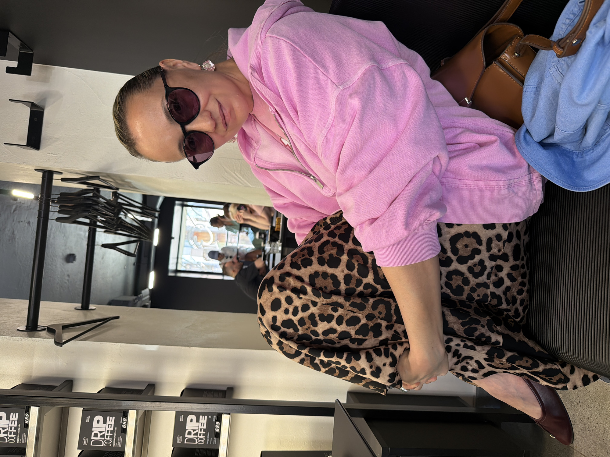 The image shows a woman seated indoors at what appears to be a cafe or coffee shop in Izhevsk, Russia. She is the main subject, looking towards the camera with a subtle smile while wearing dark sunglasses. She is dressed in a light pink zip-up hooded sweatshirt and a long, flowing leopard print skirt or dress. Her accessories include large, decorative earrings and reddish-brown flat shoes. Beside her, a brown leather handbag is visible, with a light blue denim garment draped over it.
In the background, other people are partially visible, out of focus, suggesting a public setting where people are congregating. The interior features modern design elements, including black metal shelving units or racks with hangers, and light-colored walls. Natural light enters through a window in the upper background, indicating it is daytime.
A notable detail in the foreground is a display shelf stocked with black boxes labeled "DRIP COFFEE." The text "ИНДОНЕЗИЯ СУМАТРА ГАЙО" (Indonesia Sumatra Gayo) and the number "689" are also clearly visible in Cyrillic script on these coffee boxes. The woman appears relaxed, sitting comfortably in what seems to be a casual, contemporary coffee establishment.