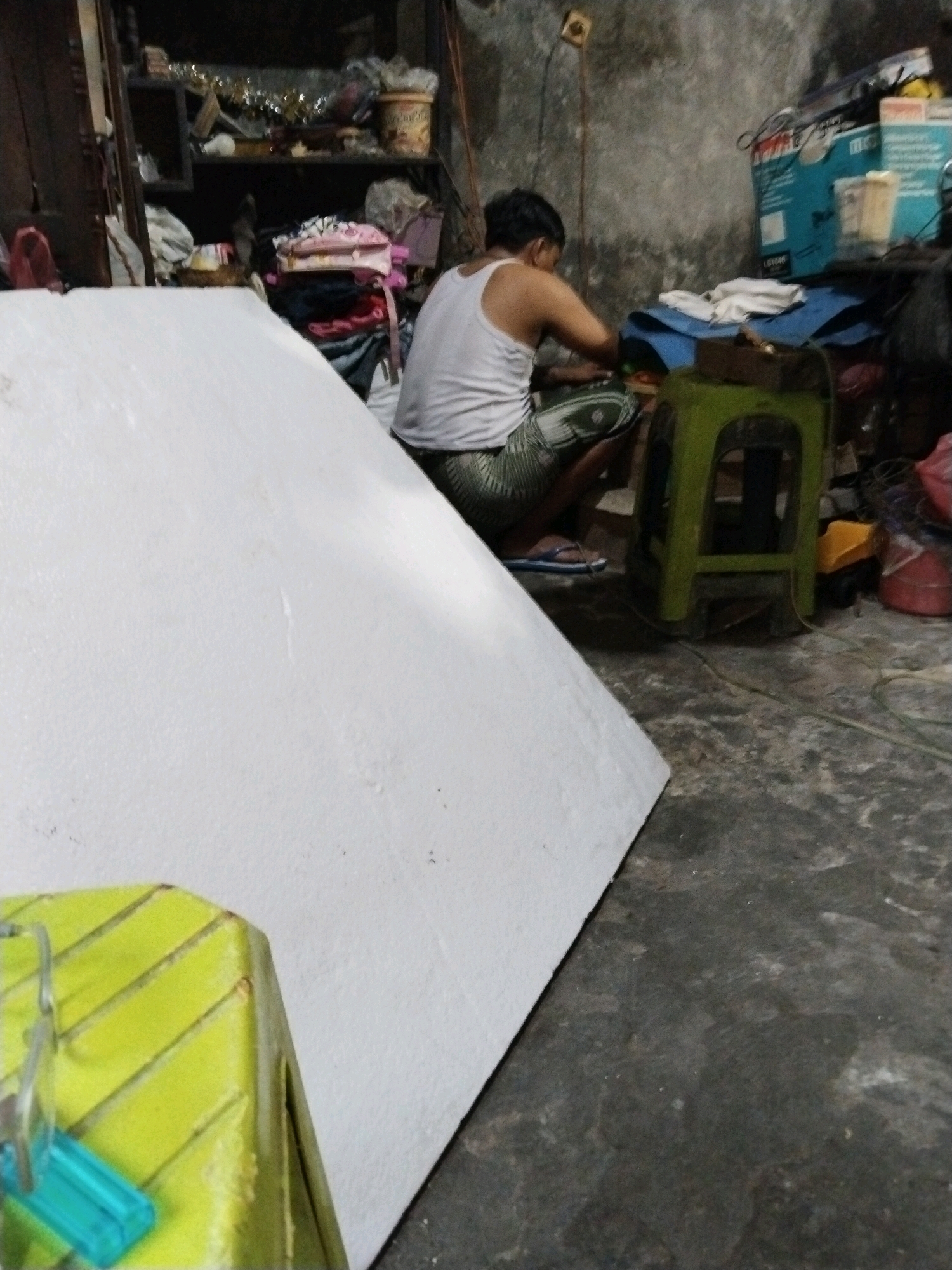 A person wearing a white tank top and patterned shorts is squatting next to a large white object. They are working with their hands on something in front of them, which is on a green stool. The environment appears to be an indoor workspace with shelves stacked with various items, including boxes, bags, and containers. The floor is concrete, and there are electrical cords visible. The overall impression is of a cluttered but functional area. The location is identified as Pepelegi, Indonesia, though no specific landmarks or street views are depicted.