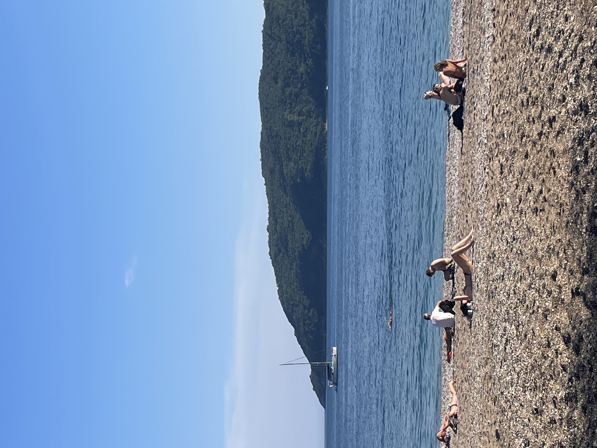 The image shows a coastal scene in Budva, Montenegro, featuring a pebble beach along the Adriatic Sea. Several people are sunbathing or relaxing on the shore. In the water, one person is swimming. A sailboat is anchored offshore, with a wooded peninsula rising in the background. The sky is clear and blue.