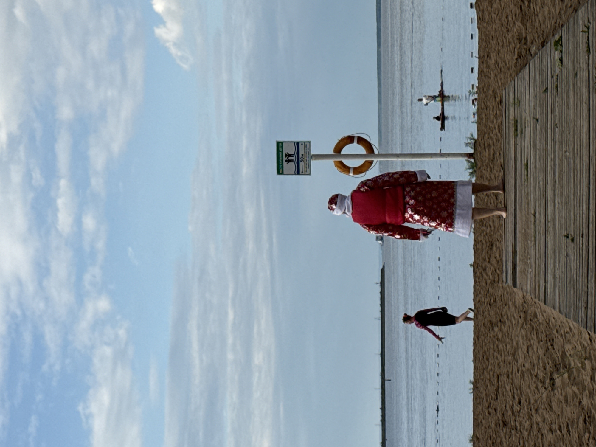 A person in a Santa Claus costume stands on a wooden boardwalk facing away from the camera on a beach. The costume is red with white trim and a patterned design. The person is barefoot. To the left, a woman in a dark swimsuit is walking on the sand towards the water. In the distance, a man and a child are on a paddleboard in the water. A sign with a lifebuoy attached to its pole is visible near the Santa Claus figure, indicating a swimming area. The background is a body of water, likely the Kama River, with a distant shoreline and a cloudy blue sky. This scene appears to be at a beach in the city of Chaikovsky, Russia, given the distinctive signage and the geographical context of the Kama River.
