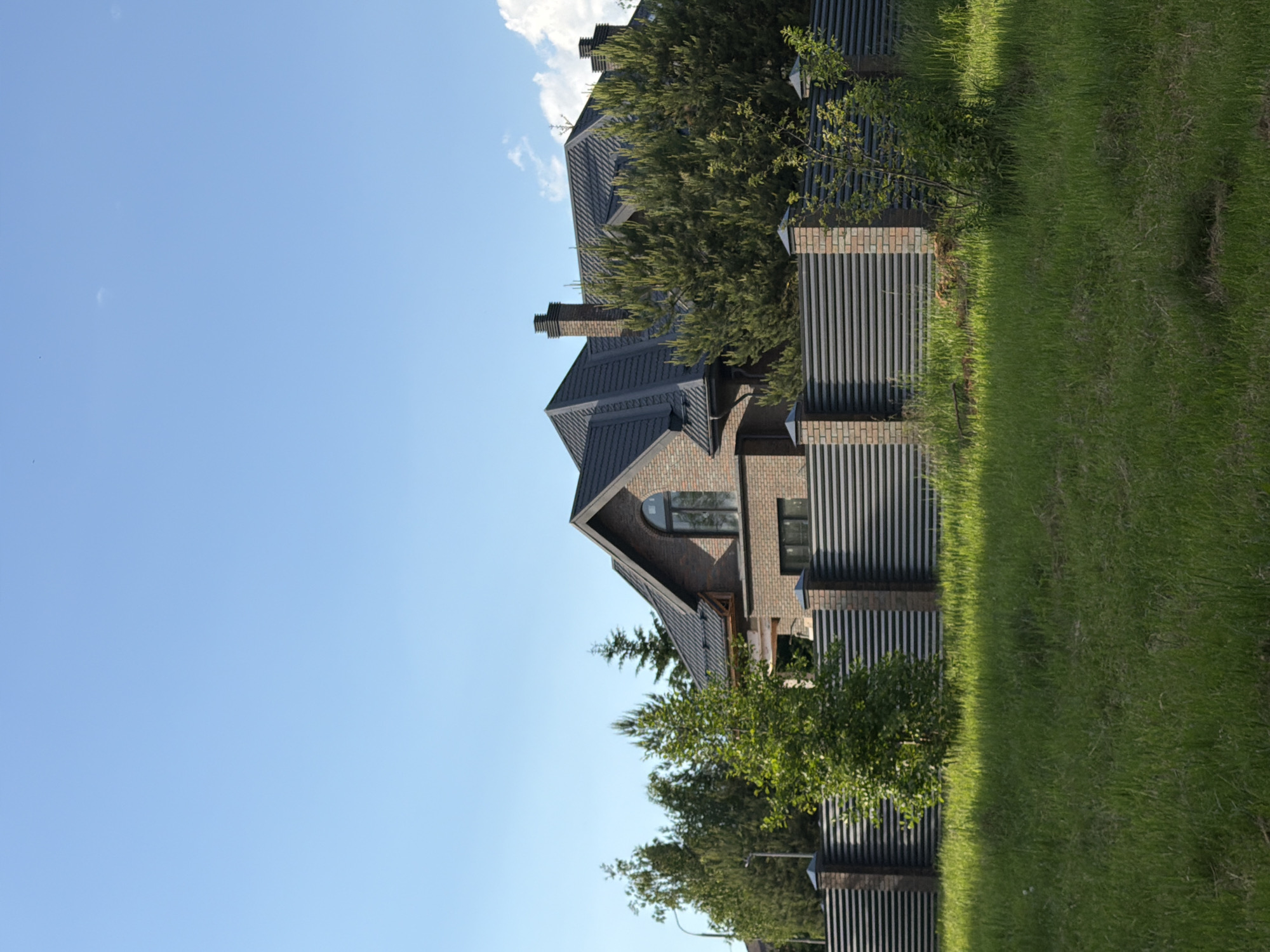 This image depicts a large, modern private residence set within a verdant landscape, likely in a residential area of Satino-Russkoe, Russia. The central subject is a multi-story house constructed primarily of dark-toned brick, possibly red or brown. Its architectural design is complex, featuring a dark-colored, multi-pitched roof with numerous gables and at least two prominent brick chimneys. A distinctive large arched window is visible on one of the upper gables, adding to the house's contemporary aesthetic, alongside other rectangular windows.
The property is delineated by a sleek, slatted fence, likely made of metal or dark wood, that runs along the foreground and sides of the house. This fence is intermittently supported by brick pillars that complement the house's exterior. The ground is covered with lush, vibrant green grass, indicating a well-maintained lawn. Various trees and shrubs are planted around the house and along the fence line, contributing to the natural surroundings and partially obscuring parts of the building.
The scene is bathed in bright, clear daylight under a light blue sky, which contains only a few faint, wispy white clouds in the upper right quadrant. This suggests a sunny day, likely morning or afternoon. No people are visible, and no specific activities are taking place, presenting a serene and static view of the residential property. The overall impression is one of modern design, order, and tranquility within a well-manicured setting. There is no visible text within the image.