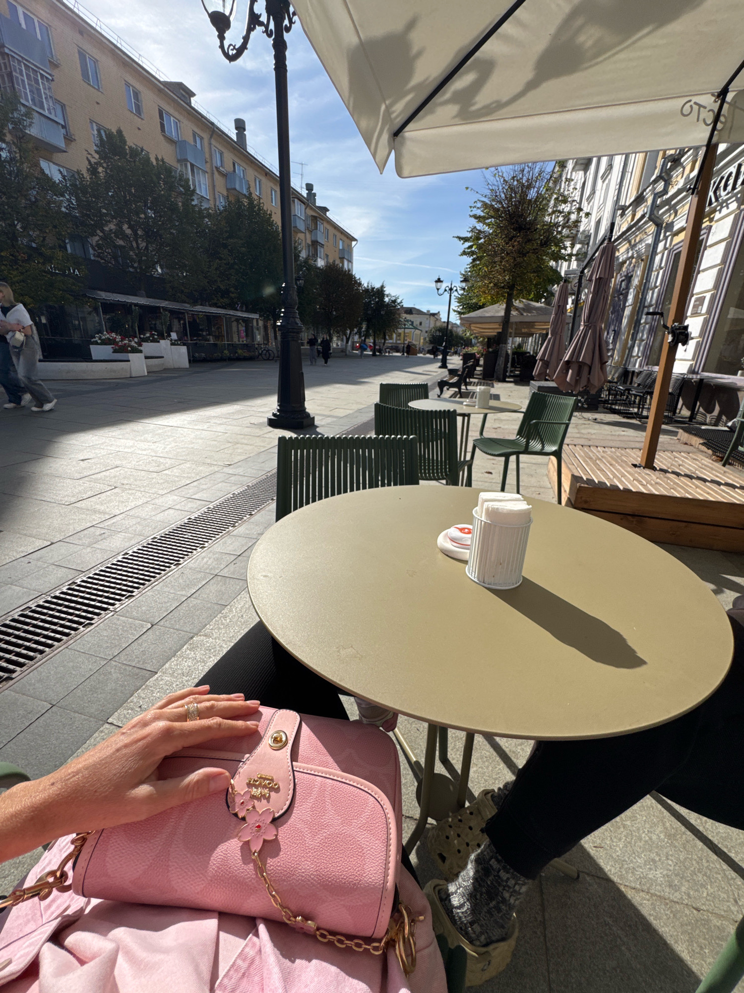 The image depicts a street scene in Tver, Russia, likely a pedestrian zone or sidewalk cafe area. In the foreground, a person's hand with a ring is resting on a pink handbag. The handbag is adorned with a gold chain and a pink cherry blossom charm. The person is wearing black pants and clogs with patterned socks. A round, light-colored table is in the center of the frame, with a napkin holder containing white napkins and a small object on it. In the background, there are cafe tables and chairs, some with umbrellas. Buildings line the street, with trees and a lamppost visible. The lighting suggests it is daytime with bright sunlight casting shadows. The overall environment appears to be an urban setting with an outdoor seating area for a cafe or restaurant.