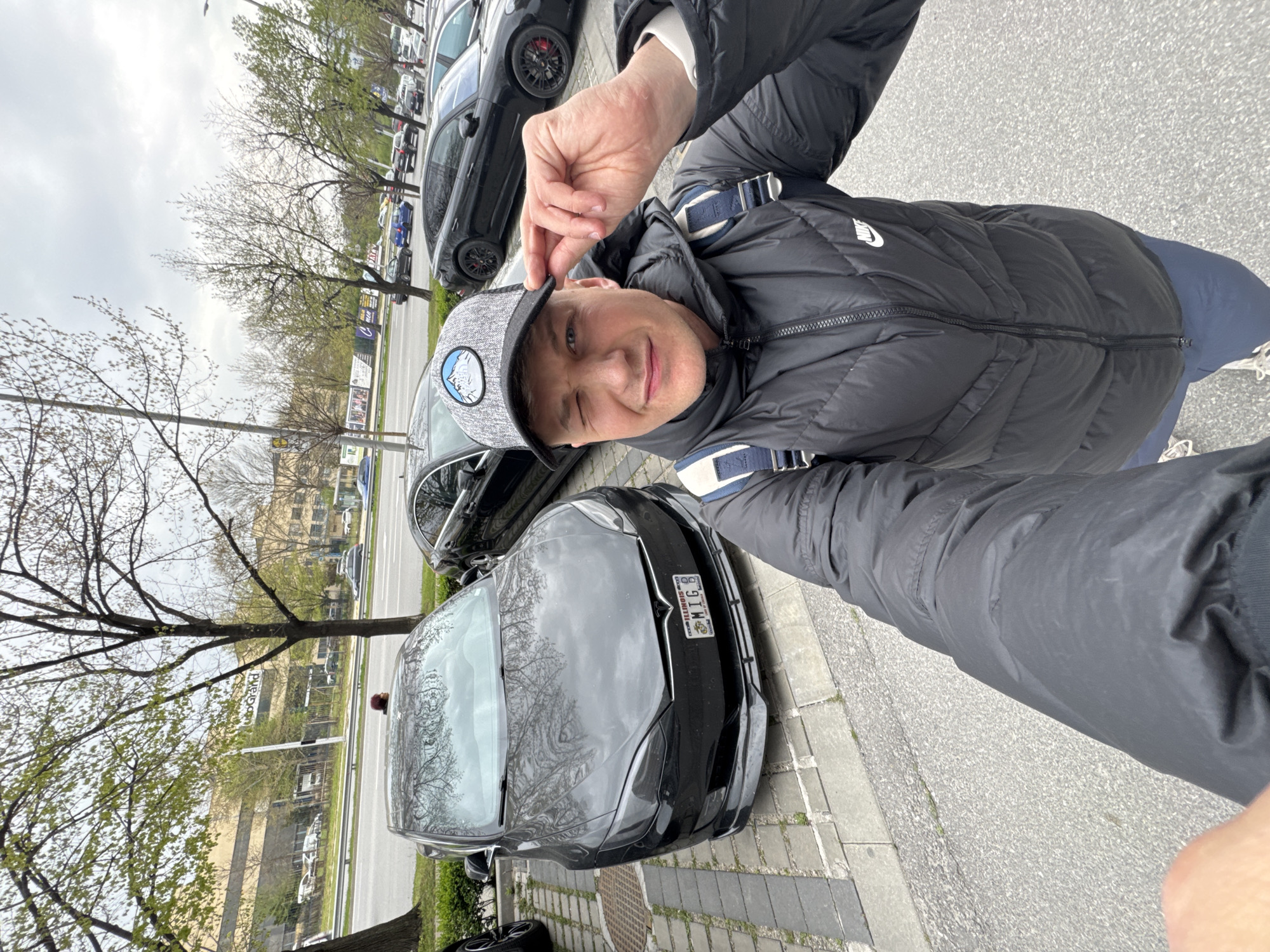 The image shows a man taking a selfie. He's wearing a black puffer jacket with a white Nike logo on the sleeve, a grey patterned baseball cap with a circular logo, and blue trousers. He's winking and touching his cap with his right hand.
In the background, there's a black Tesla car parked on a paved area. The car has an Illinois license plate that reads "MIC 6". Behind the Tesla, there's another black car, likely a Porsche.
The scene appears to be a street with trees and buildings in the background. There are several other parked cars visible in the distance. The sky is overcast. The scene is located in Plovdiv, Bulgaria. There is a building with the sign "Puhrand" on it in the background.