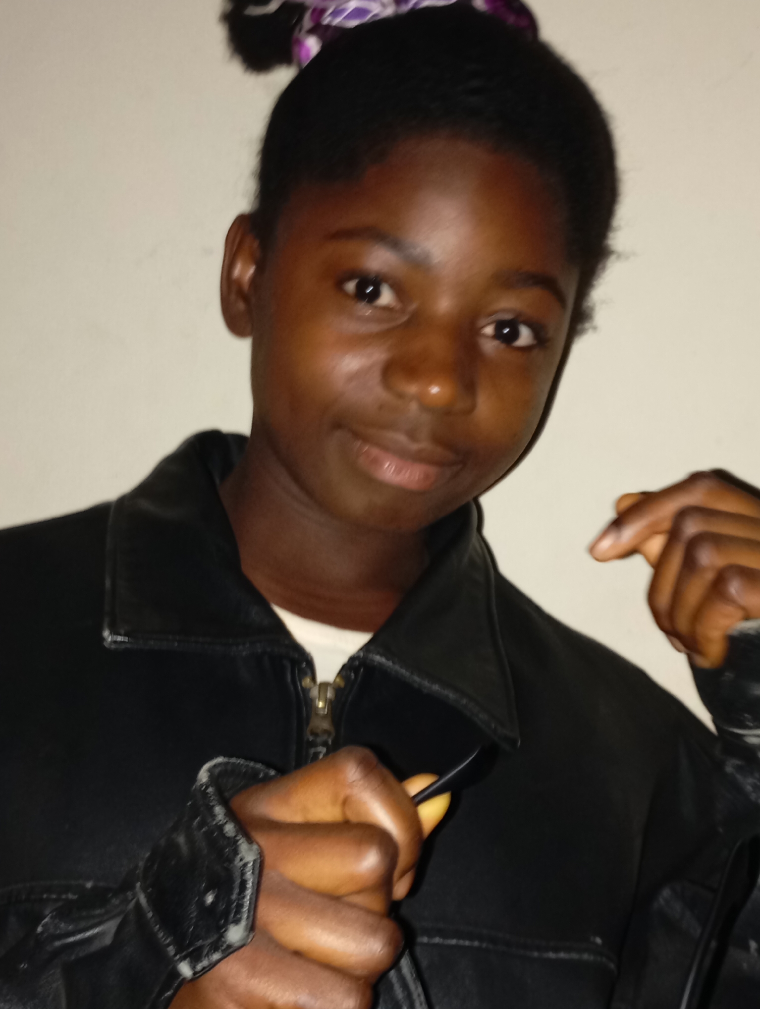 A young, dark-skinned woman with her hair pulled back, possibly in a bun, is posing for a photo in what appears to be an indoor setting. She is wearing a black leather jacket over a white top, partially zipped up. Her hands are partially visible in the foreground. The background is a plain, off-white wall. There is nothing in the scene indicating a specific location in Jos, Nigeria.