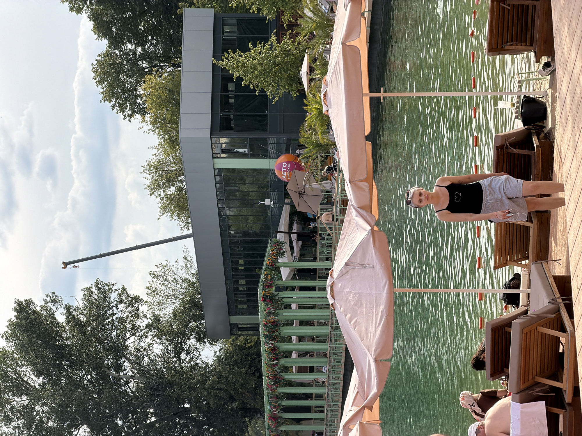 This image depicts a lively outdoor scene at a beach club in Moscow, Russia. In the foreground, a young woman with blonde hair and sunglasses is walking on a wooden deck towards the camera. She is wearing a black tank top with a heart emblem and gray shorts. Behind her, several lounge chairs are set up, some occupied by people relaxing by the water. The water itself appears to be a pool or a man-made body of water, with orange buoys marking a boundary.
In the mid-ground, a long, green structure adorned with colorful flowers runs parallel to the water. People can be seen seated at tables under this structure. Further back, a modern, multi-story building with large glass windows stands prominently. An orange balloon with Cyrillic text that appears to read "ЛЕТО В МОСКВЕ" (Summer in Moscow) is visible near the building. Above the building, a construction crane extends into the cloudy sky, indicating ongoing development or renovation. The overall atmosphere suggests a recreational area, likely a popular spot for leisure and entertainment during warmer months in Moscow.