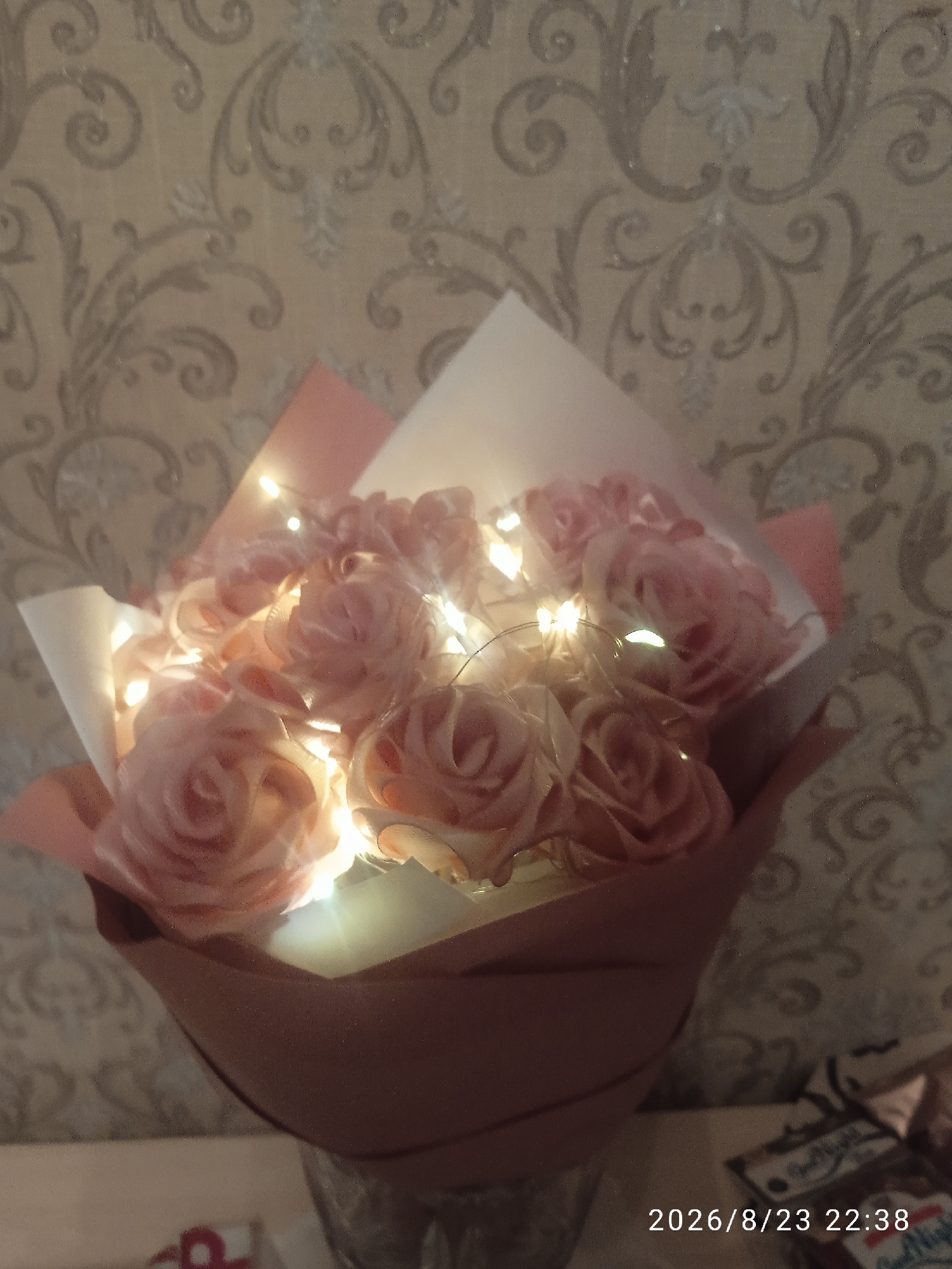 A close-up shot focuses on a bouquet of pink artificial roses, interspersed with small, warm-toned LED lights. The roses are arranged in a slightly tilted manner, with a translucent white paper cone and pink paper wrapping at the base. Behind the bouquet, a textured beige wallpaper with a raised, ornate damask pattern is visible. The lighting suggests an indoor setting, likely at night, given the timestamp "2026/8/23 22:38" which appears in white text at the bottom right of the image. A blurry, out-of-focus object, possibly a book or packaging with some text, is visible in the lower right corner, partially obscuring the timestamp. The overall scene conveys a sense of decoration or a gift, possibly for a special occasion. The city of Lida, Belarus is not depicted in this image.