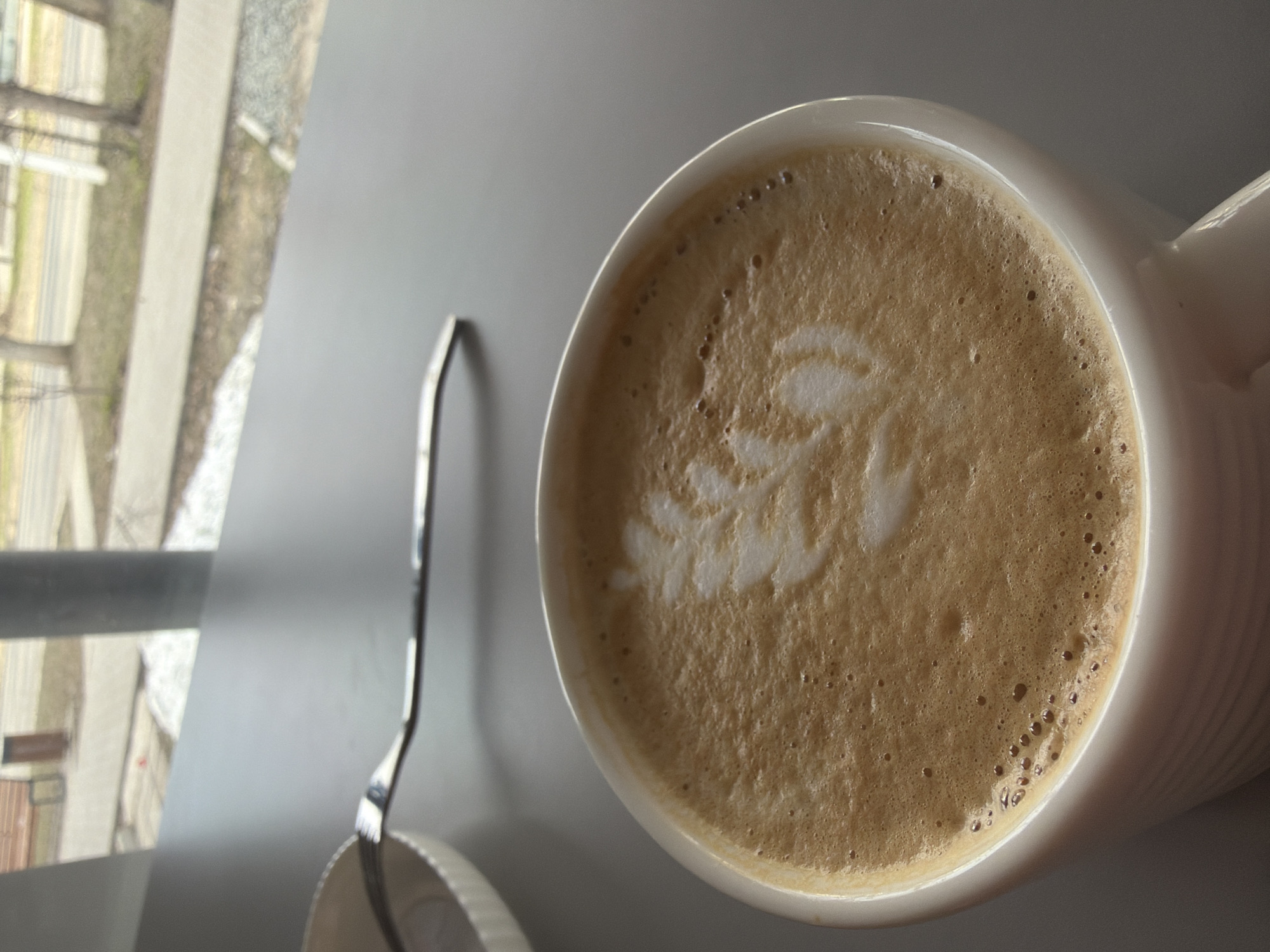 An overhead photograph captures a white ceramic mug filled with coffee, likely a latte or cappuccino, resting on a smooth, grey table or countertop. The coffee features intricate latte art, resembling a stylized white leaf or feather pattern, set against a frothy, light brown surface with small bubbles. The mug itself is white and has subtle vertical ridges on its visible side.
To the left of the mug, a slim, metallic spoon is partially visible, suggesting it's ready for use. Further to the left, a large window offers a blurred view of an outdoor environment. This exterior scene depicts what appears to be a paved path or street, flanked by bare trees or sparse vegetation, and indistinct building outlines in the distance. The soft, natural light entering from the window indicates that the photograph was taken during the daytime.
The setting appears to be indoors, looking out onto an urban or suburban landscape in Chaikovsky, Russia. The blurry background emphasizes the warm, inviting presence of the coffee indoors. There are no people visible, and no discernible text is present anywhere in the image. The overall mood conveyed is one of tranquility and comfort, capturing a quiet moment of refreshment.