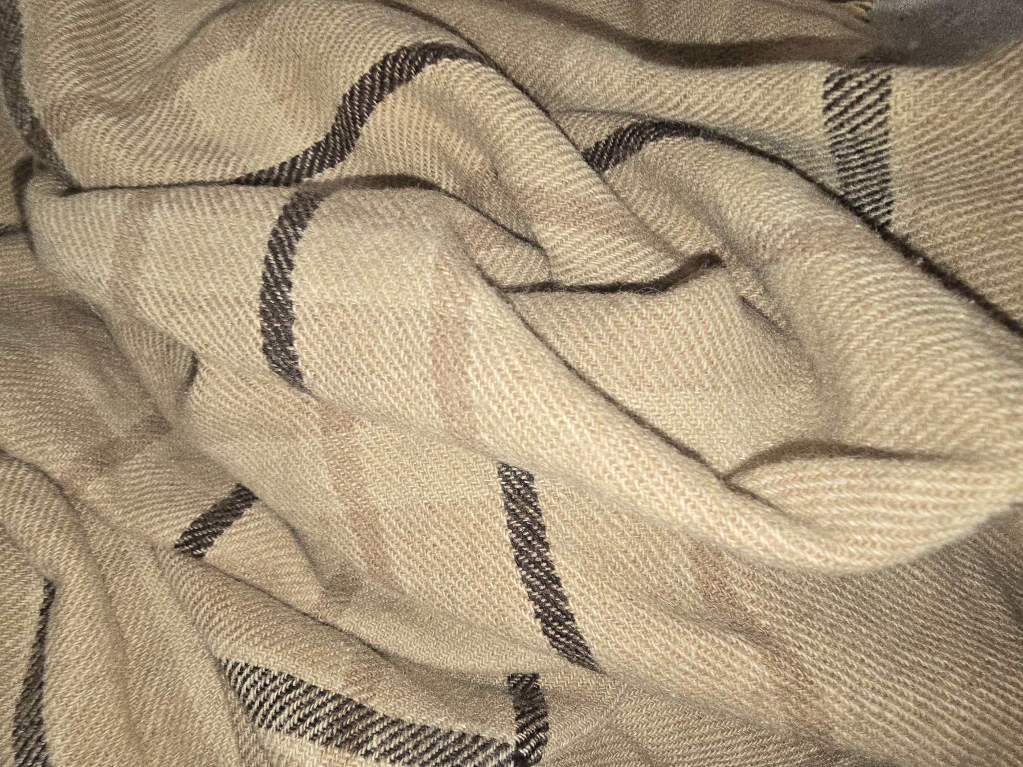 The image shows a close-up, detailed view of a light brown plaid blanket. The blanket has a textured weave and features dark brown and black stripes arranged in a tartan pattern. The fabric is folded and draped, creating shadows and highlights that emphasize the texture and the crisscrossing lines of the plaid. There are no people or any other objects visible in the image, and no environmental context is provided. The image does not contain any information that would allow for its location to be identified, either within the city of Ulan-Ude, Russia, or anywhere else.
