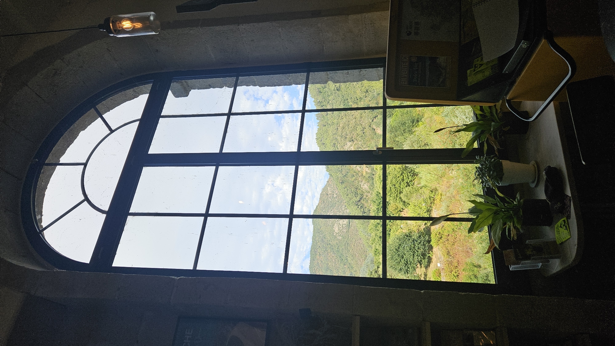 The image depicts a view from inside a building looking out through a large, arched, multi-paned window. The window overlooks a lush, green landscape of hills and trees under a bright, partly cloudy sky. In the foreground, on a table inside the building, are several potted plants, some small brochures or leaflets, and a dark, possibly stone, object. To the right, a portion of a yellow display stand or cabinet is visible, with some papers or publications on it. A hanging light fixture with an exposed bulb is present in the upper right corner. The architecture visible around the window suggests a rustic or historical setting, possibly a stone building. The location is identified as Saint-Sauveur-de-Montagut, France.