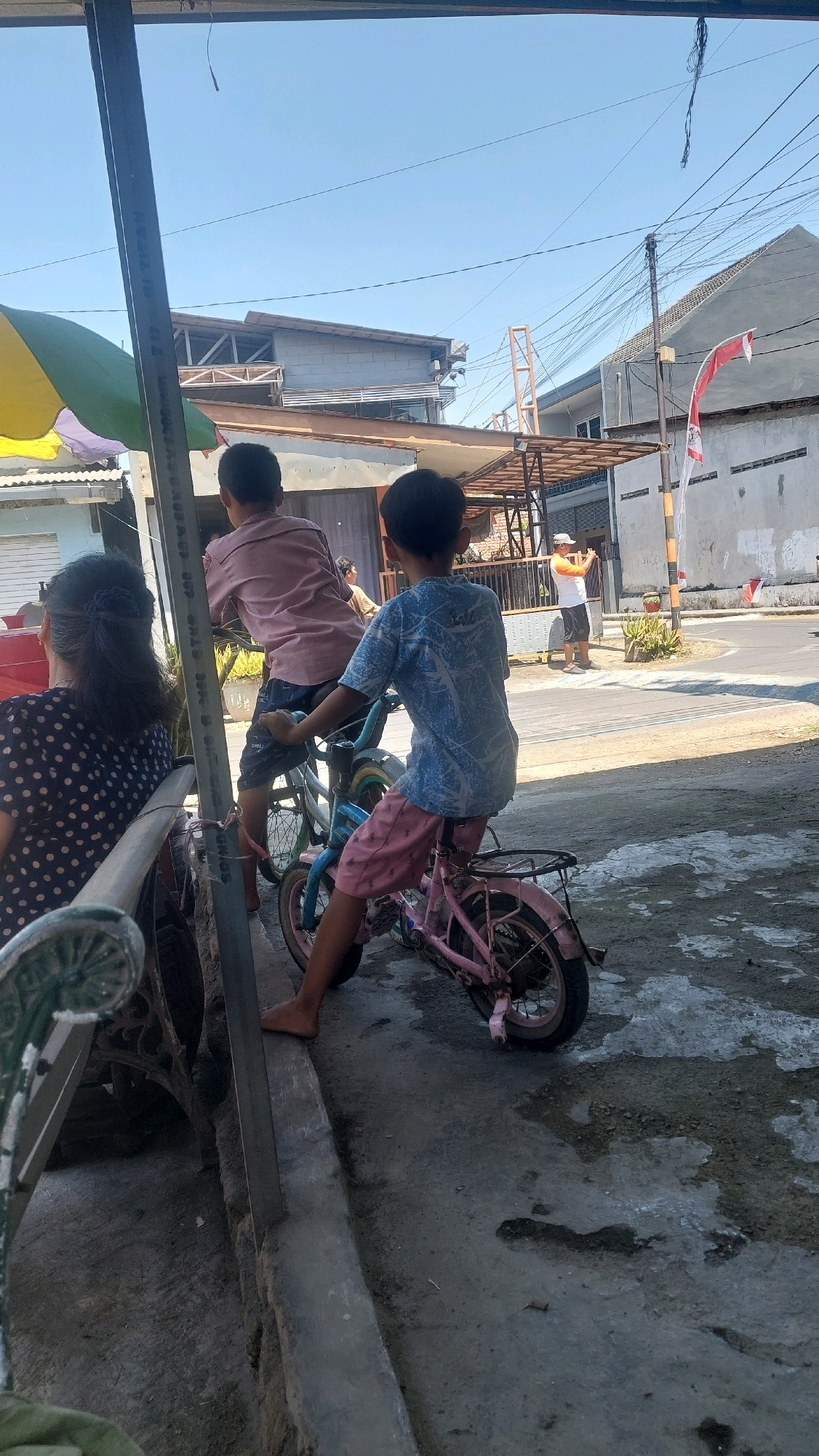 In Sidoarjo, Indonesia, two young boys are seen riding bicycles on a street. The boy in the foreground is on a pink bicycle and wears a blue t-shirt and pink shorts. The boy behind him is on a blue bicycle and wears a pink shirt and dark shorts. To the left of the frame, a woman with dark hair, wearing a black and white polka dot dress, sits on a bench. The street is paved with asphalt and concrete, with a concrete curb running along the side. In the background, buildings with various roofing styles and utility poles with numerous wires are visible under a clear blue sky. A man in an orange and white shirt and a cap is standing in the distance, holding a phone to his ear. A red and white flag is attached to a pole on the right side of the image. The scene appears to be a casual outdoor setting in a residential or commercial area.
