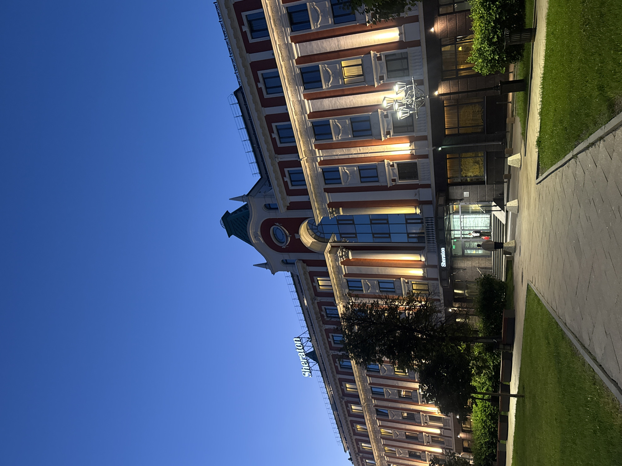 The image shows the Sheraton hotel in Nizhny Novgorod, Russia, at dusk. The hotel is a multi-story building with a red and white facade, illuminated by exterior lights. A prominent "Sheraton" sign is visible on the rooftop. In front of the hotel, there is a paved walkway, a grassy area with benches, and trees. Two people are walking towards the entrance of the hotel. The sky is a clear, deep blue.