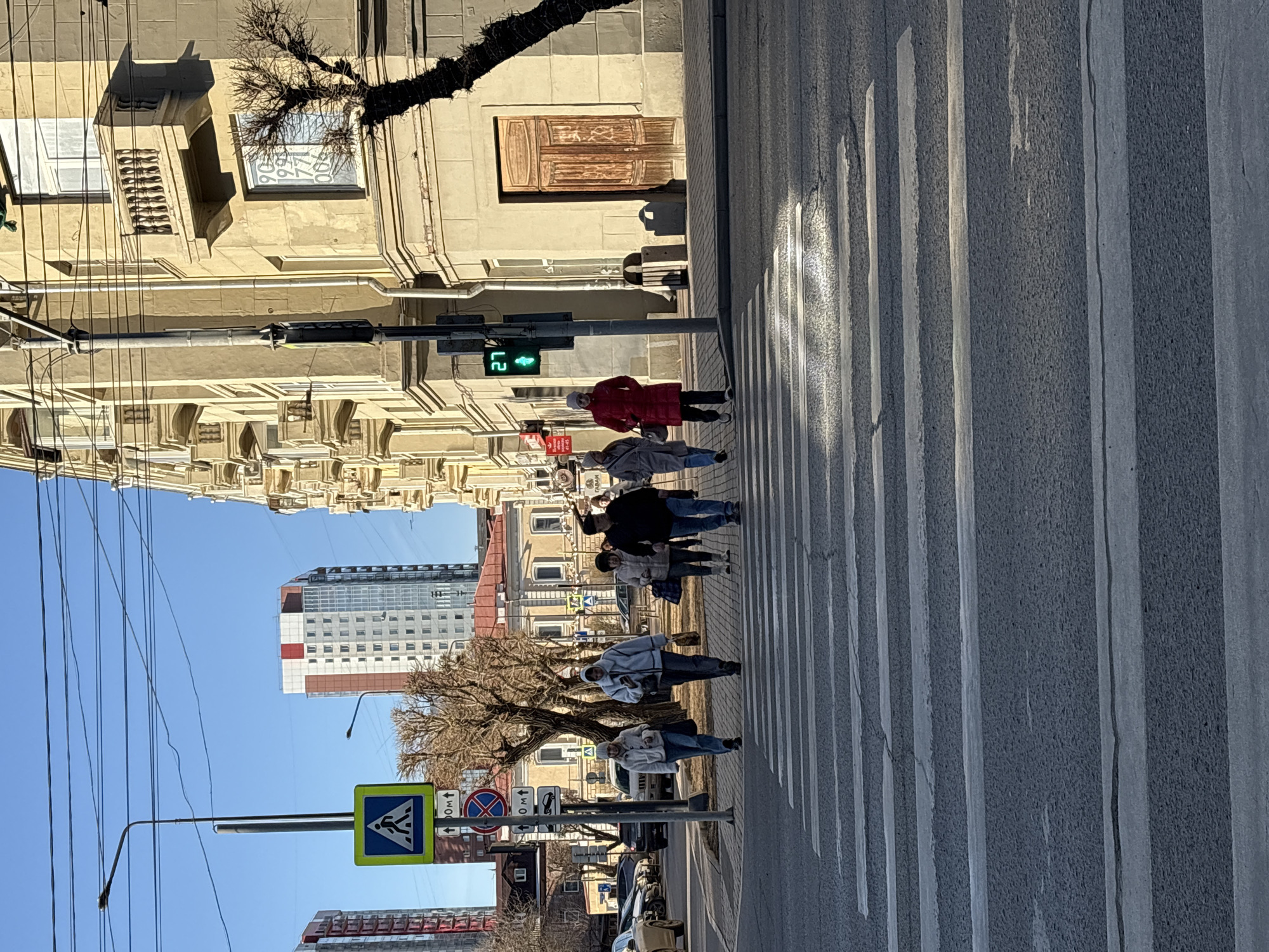 The image depicts a group of pedestrians crossing a street at a crosswalk in Krasnoyarsk, Russia. A traffic light shows a green walking signal. The crosswalk has thick white stripes on asphalt. A building with an ornate facade is on the left, with a clock on the upper part of the building. A modern building is visible in the background. A street sign for a pedestrian crossing is visible. Cars are parked along the street. Overhead power lines are present. A bare tree stands on the street. Pedestrians are wearing a variety of clothing, including a red coat.