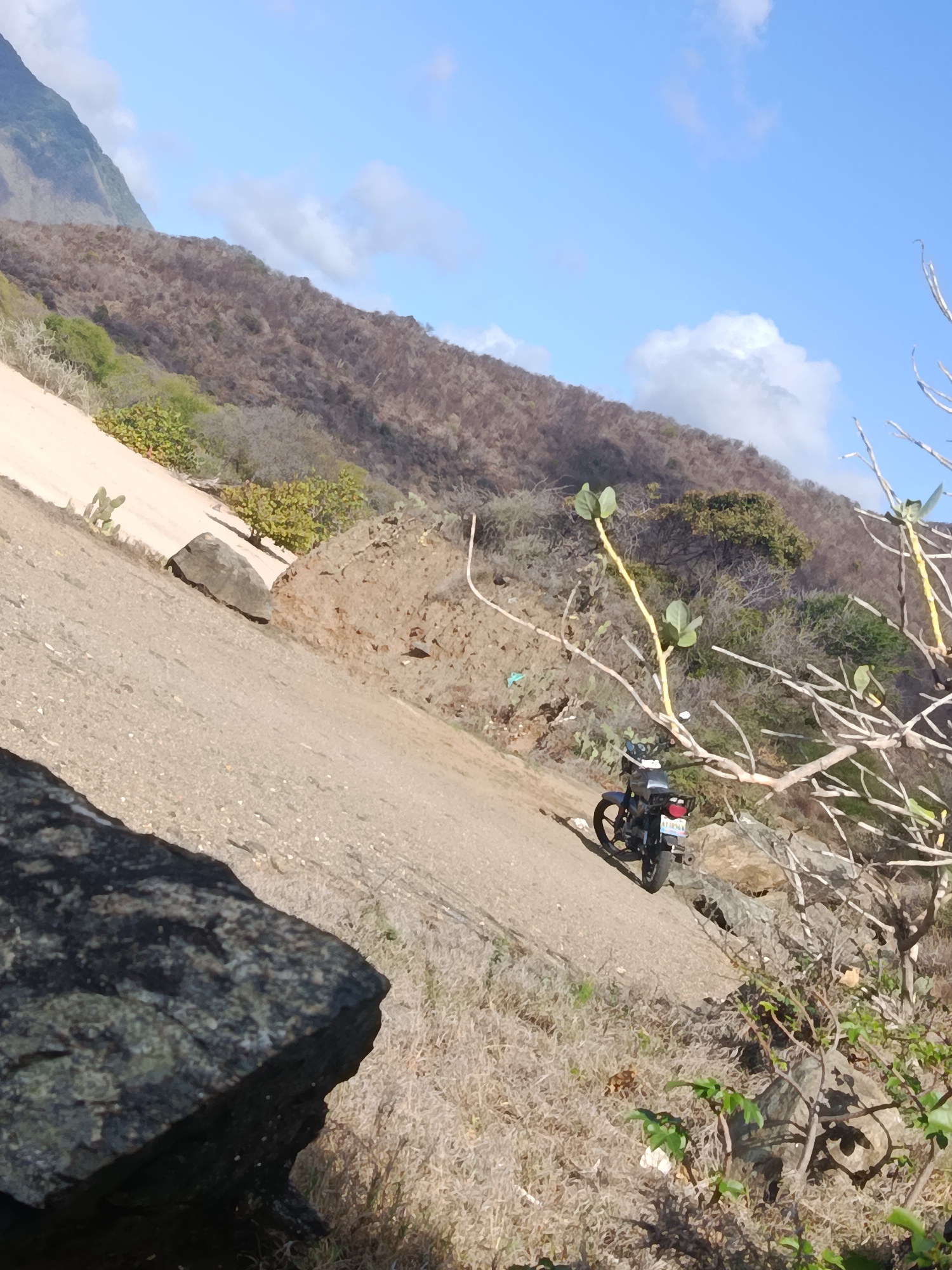The image depicts a dry, mountainous landscape in Pedro González, Venezuela. In the foreground, a large dark rock is visible on the left side, with dry grass and scrub vegetation. A dirt path slopes upwards diagonally across the frame. On the path, a black motorcycle with a light-colored license plate is parked. In the middle ground, the terrain is rocky and barren, with scattered bushes and some sparse vegetation. Further up the slope, there is a lighter-colored sandy area with a few small green bushes and cacti. The background is dominated by a dry, brown mountainside with sparse trees and vegetation, leading up to a hazy blue sky with some white clouds. The overall impression is one of a rugged, arid environment.