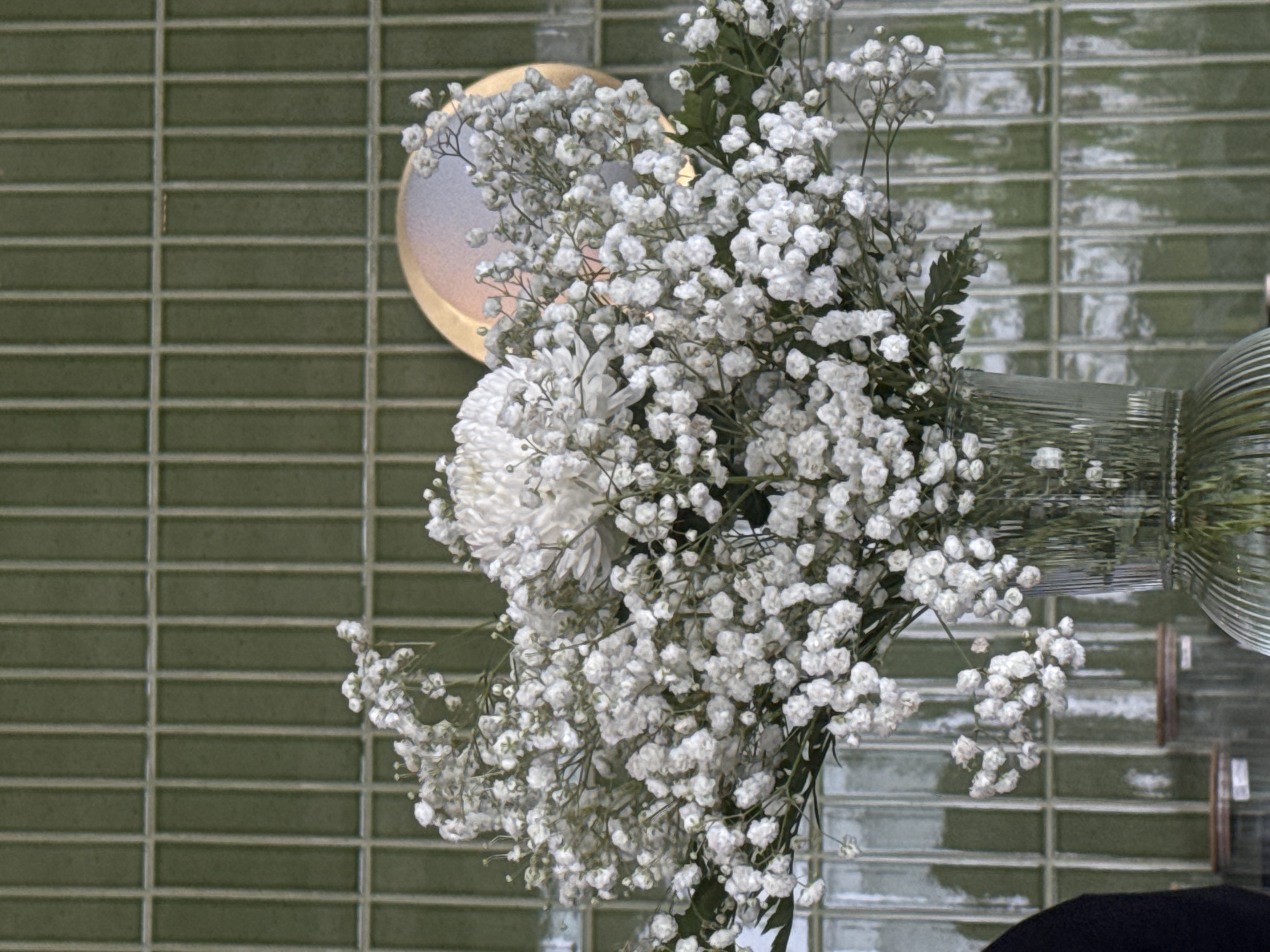 An overhead, medium shot captures a bouquet of white flowers arranged in a clear glass vase. The bouquet features a large, white chrysanthemum at its center, surrounded by a dense cluster of baby's breath. The flowers are positioned in the foreground, with the vase resting on a surface that is mostly obscured. The background consists of a wall covered in green, rectangular tiles arranged vertically. To the right of the bouquet, a circular, golden-hued object with a reflective surface is partially visible. The scene appears to be indoors, with no discernible outdoor elements.
