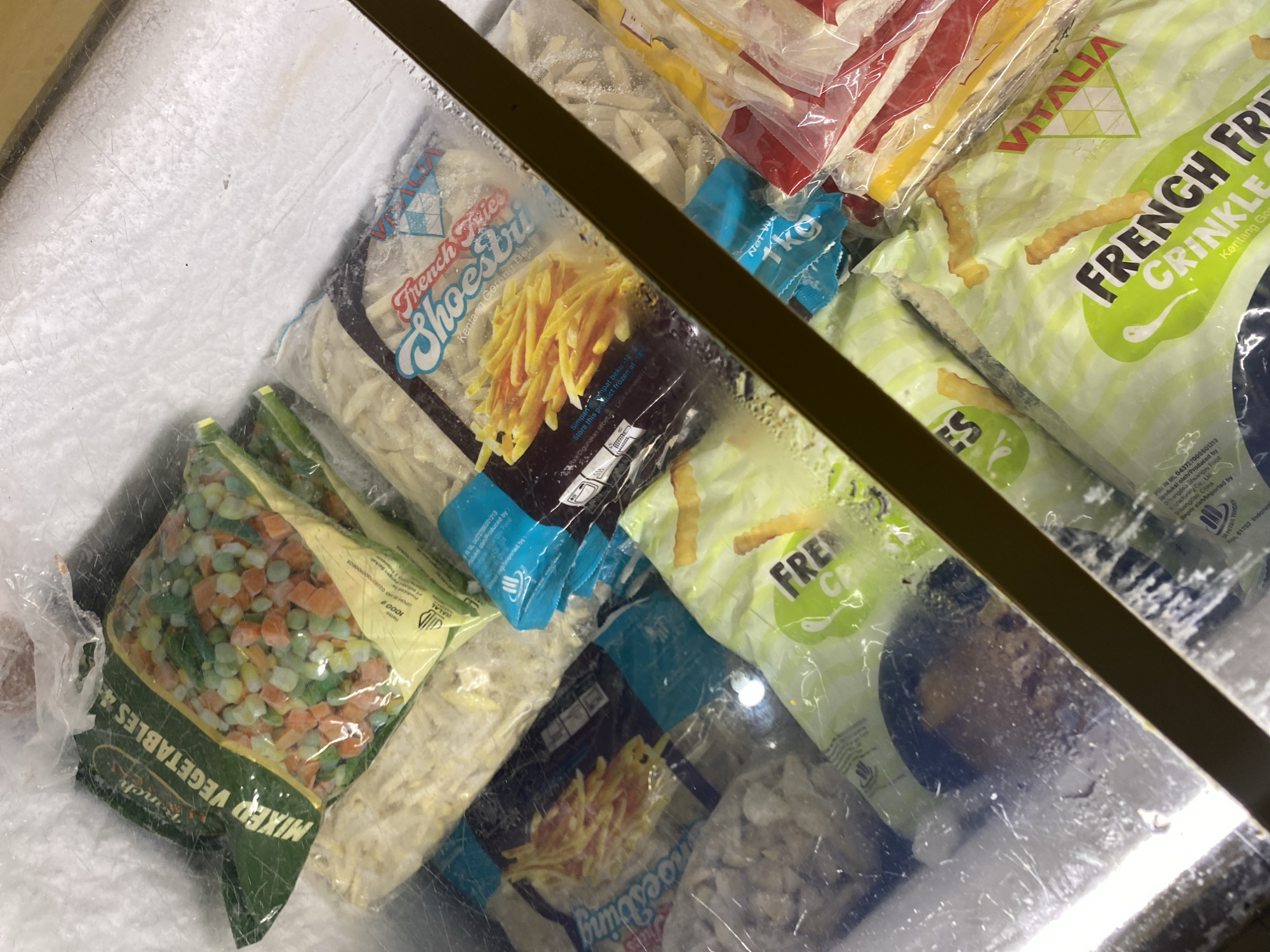 The image displays a freezer filled with various frozen food items, primarily bags of French fries and mixed vegetables. The packaging suggests a retail or food service environment, with clear labels indicating brands and product types. One prominent bag features "French Fries Crinkle" and another shows "French Fries Shoestrings." A bag of "Mixed Vegetables" is also visible. The freezer is covered by a transparent lid, through which the contents are viewed. The overall impression is one of frozen goods stored for preservation. The location is stated to be in the city of Payatieng, Indonesia.