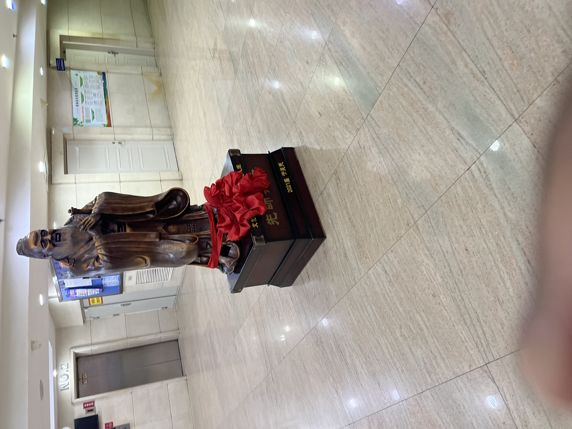 A bronze-colored statue of a man stands on a dark-colored pedestal indoors. The statue wears what appears to be traditional clothing. A red ribbon-like decoration is draped over the statue and pedestal. The pedestal has gold-colored text on its surface. The floor is tiled. In the background are doors, posters, and an elevator. Text reading "NO.2" is visible on a wall. The location is likely an academic or administrative building in Changchun City, China, based on the statue and the general interior setting.