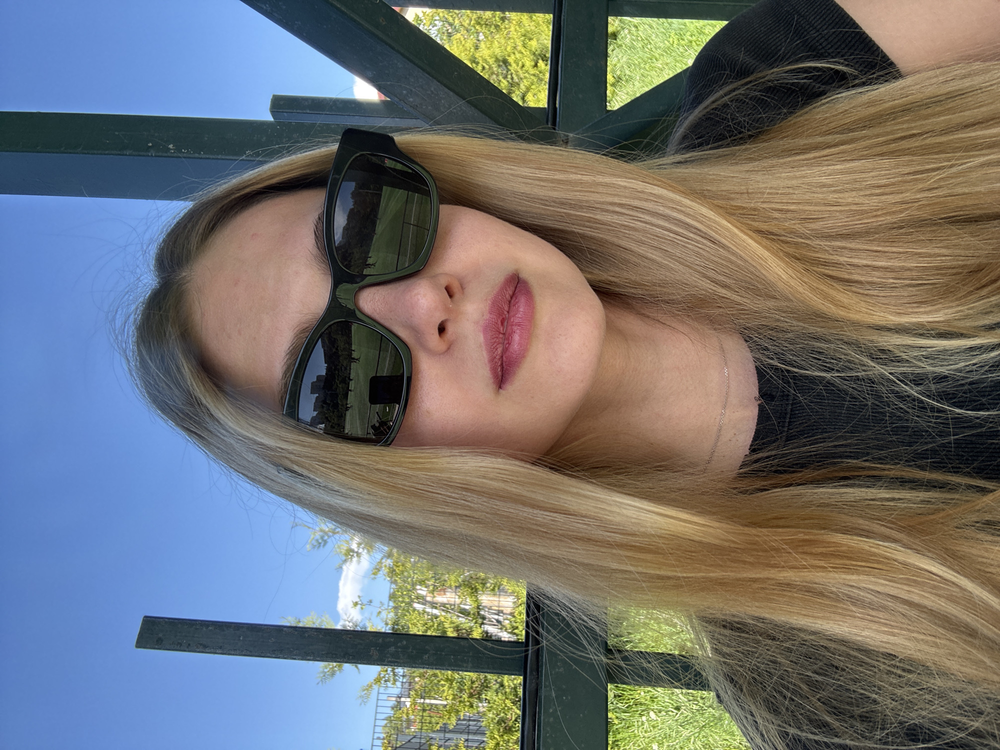 A blonde woman wearing sunglasses is reclining on what appears to be a metal park bench or similar structure outdoors. The environment includes green vegetation, a blue sky, and metal structures. The reflections in the sunglasses show buildings or architectural elements, suggesting an urban environment. Without additional contextual cues, definitively pinpointing the location to Tbilisi, Georgia, is not possible.