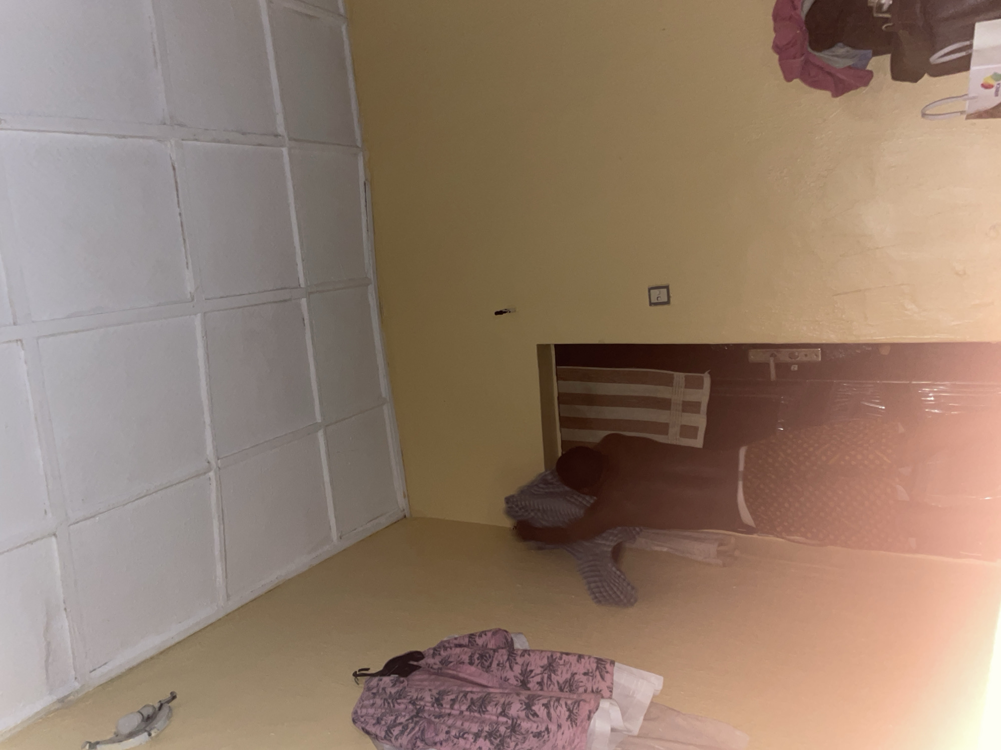 In a dimly lit room, likely in Abuja, Nigeria, a shirtless individual stands near a doorway. They are wearing patterned shorts and appear to be adjusting or holding a checkered garment. To the left, several articles of clothing are hung on the wall, including a pink and white patterned shirt. The room's walls are painted a pale yellow, and the ceiling is a white tiled surface. A light fixture is visible on the wall near the ceiling on the left side of the frame. A doorway with a vertical striped curtain hangs beyond it, and a doorknob is visible. A light switch is mounted on the wall to the right of the doorway. Further to the right, a pile of clothes, including a pink garment, sits atop a dark object. A white shopping bag with a colorful logo rests on the floor in the foreground on the right. The lighting in the room is uneven, with a strong, diffused light source coming from the lower left, creating a lens flare effect.