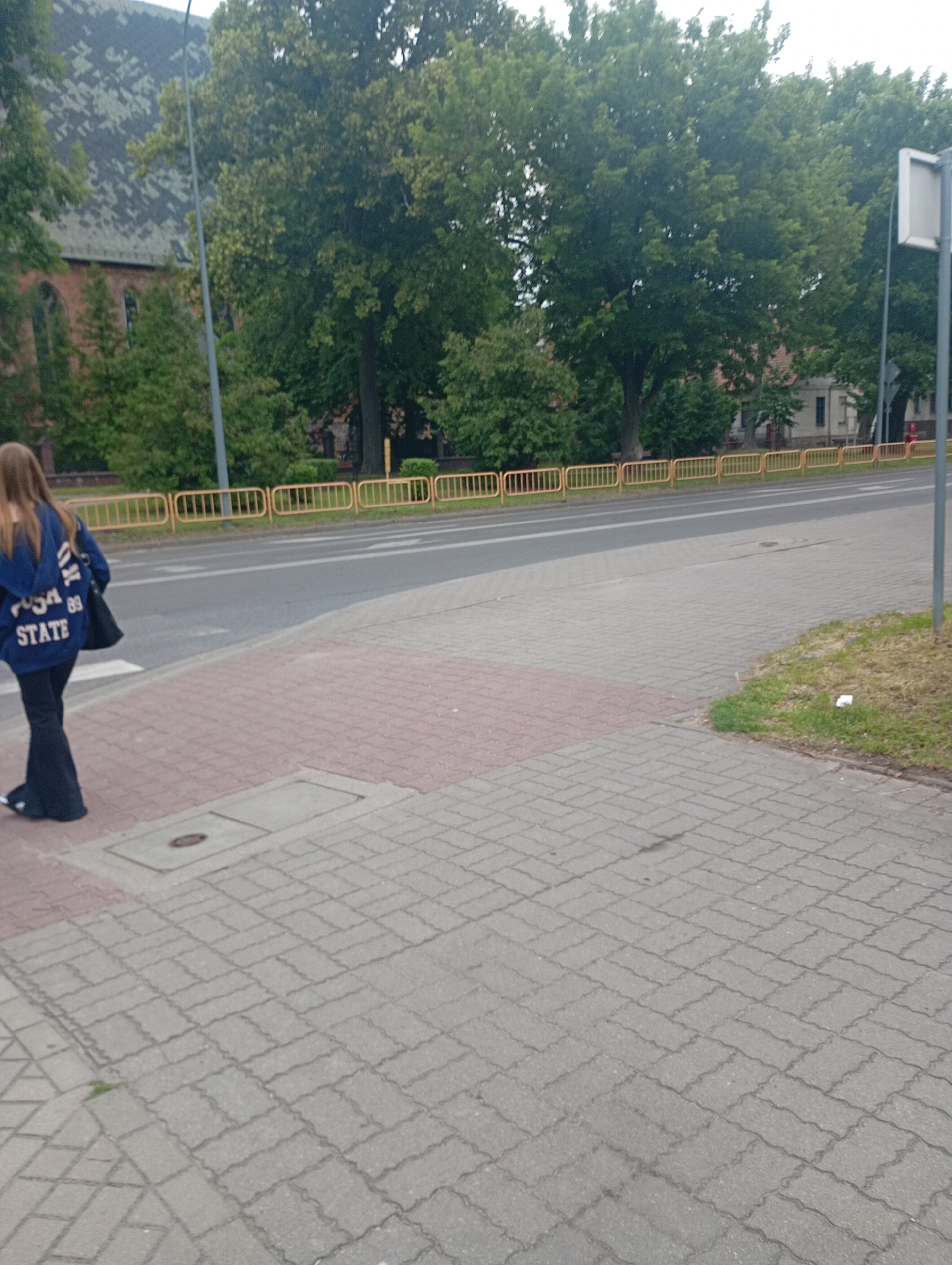 In Międzyrzecz, Poland, a woman with long, light brown hair is walking away from the camera across a pedestrian crossing. She is wearing a dark blue hooded sweatshirt with white lettering that appears to say "USA 89 STATE" and black flared pants. She carries a black shoulder bag. The pedestrian crossing is paved with red and grey interlocking bricks. Beyond the crossing is a paved sidewalk, and further on, a street with a yellow metal railing separating it from a grassy area and trees. In the background, a building with a dark, textured roof and red brick walls is visible, along with mature trees.