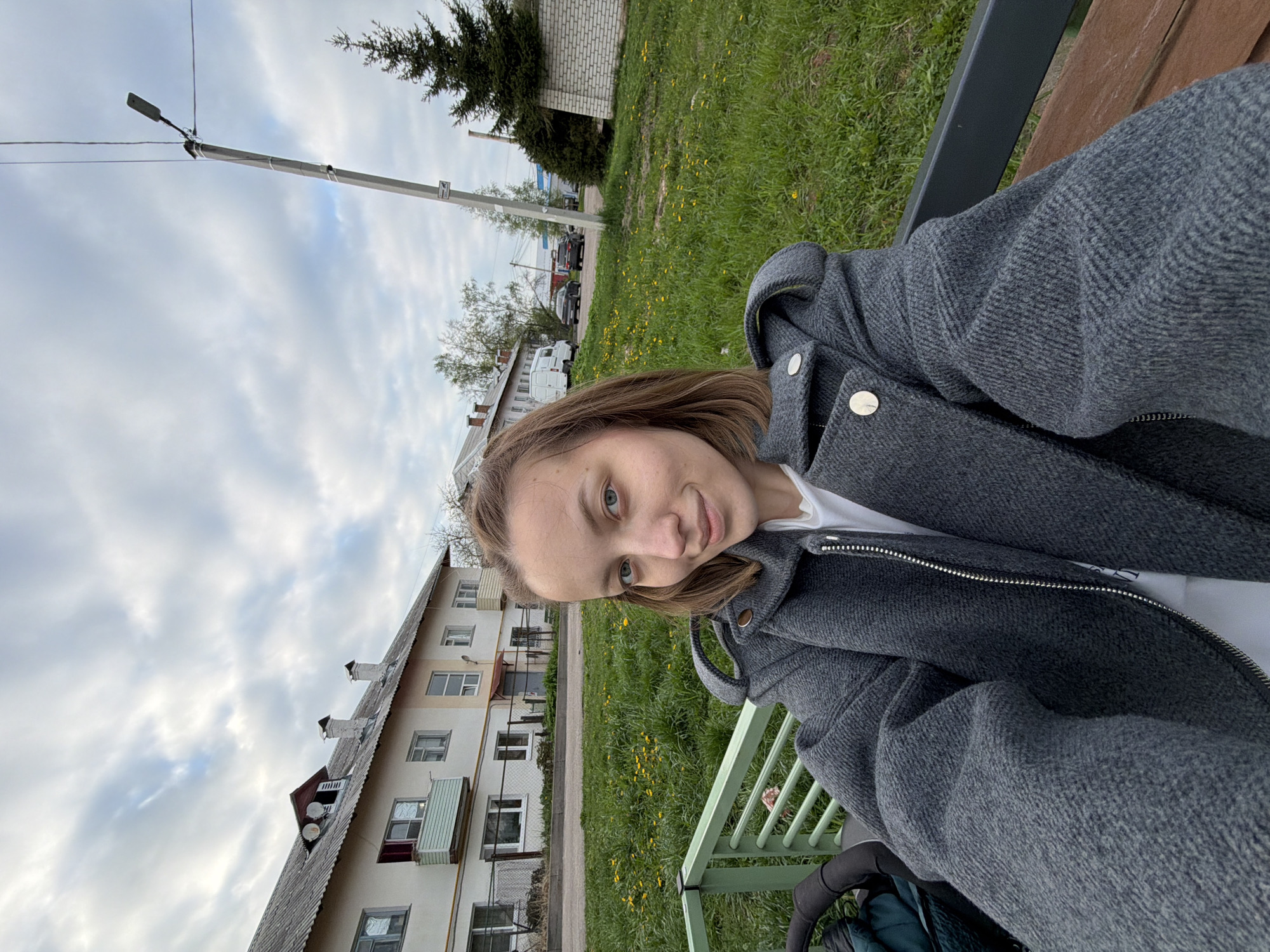 A woman sits on a green metal bench in Reshetnikovo, Russia. She is wearing a gray hooded jacket over a white shirt. Buildings are visible in the background. There is green grass and a light pole. A gray sky with clouds is overhead.