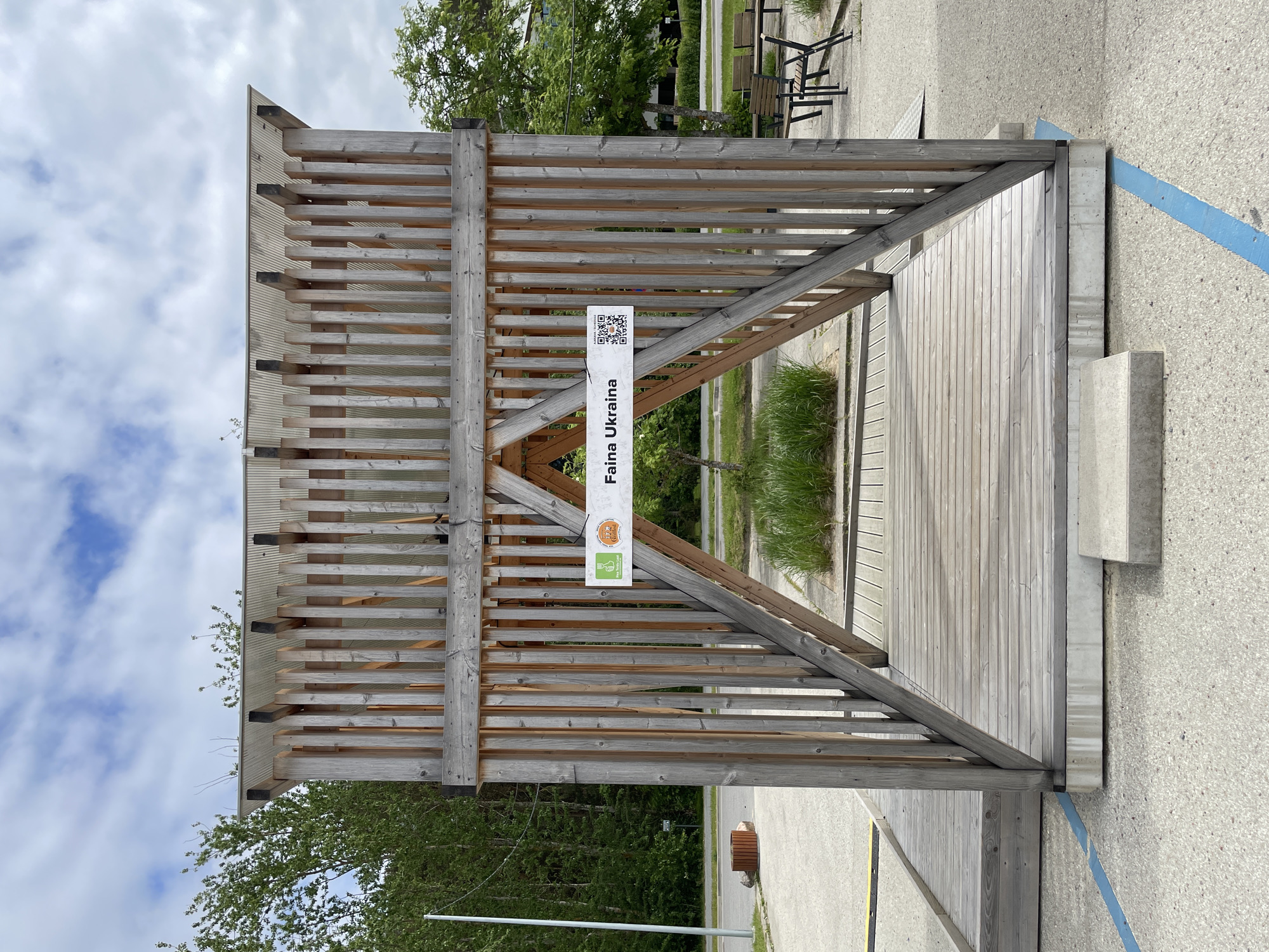 The image captures an outdoor wooden structure, potentially a pavilion or sheltered area, in Tallinn, Estonia. The structure is primarily constructed from light-colored wood, featuring a lattice-like roof and supportive beams. A sign hangs from the structure, displaying the words "Faina Ukraina" and a QR code, along with logos. The flooring of the structure is made of wooden planks. The surrounding environment includes trees, grass, benches, and paved surfaces. A blue painted line is visible on the ground near the structure, possibly indicating a parking space. The sky is cloudy, suggesting an overcast day.