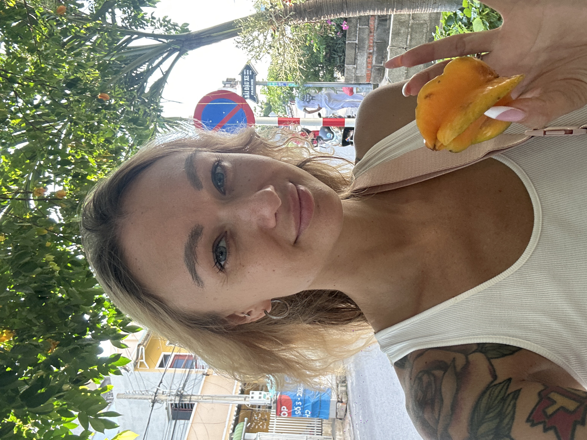 A blonde woman with blue eyes and light freckles, wearing a white ribbed tank top and a floral tattoo on her left upper arm, smiles while looking directly at the camera. She has small hoop earrings and a tragus piercing in her left ear. Her left hand, with pink and white French-tipped nails, holds a ripe yellow carambola (star fruit). The scene unfolds on an urban street in Mũi Né, Vietnam, confirmed by Vietnamese text on visible signage. Overhead utility lines are densely strung across the upper-left background. Buildings with light-colored facades, including a grey structure and a yellow building behind it, line the street. A large tree with green foliage and yellow-orange fruits, consistent with carambola, occupies the upper frame. On the right, a red and white striped pole stands next to a circular red "No Entry" sign and a white rectangular sign displaying "BÃI ĐẬU XE" (PARKING) in black Vietnamese and English text. A person in white clothing walks away in the mid-ground, near a partially visible dark-colored scooter.