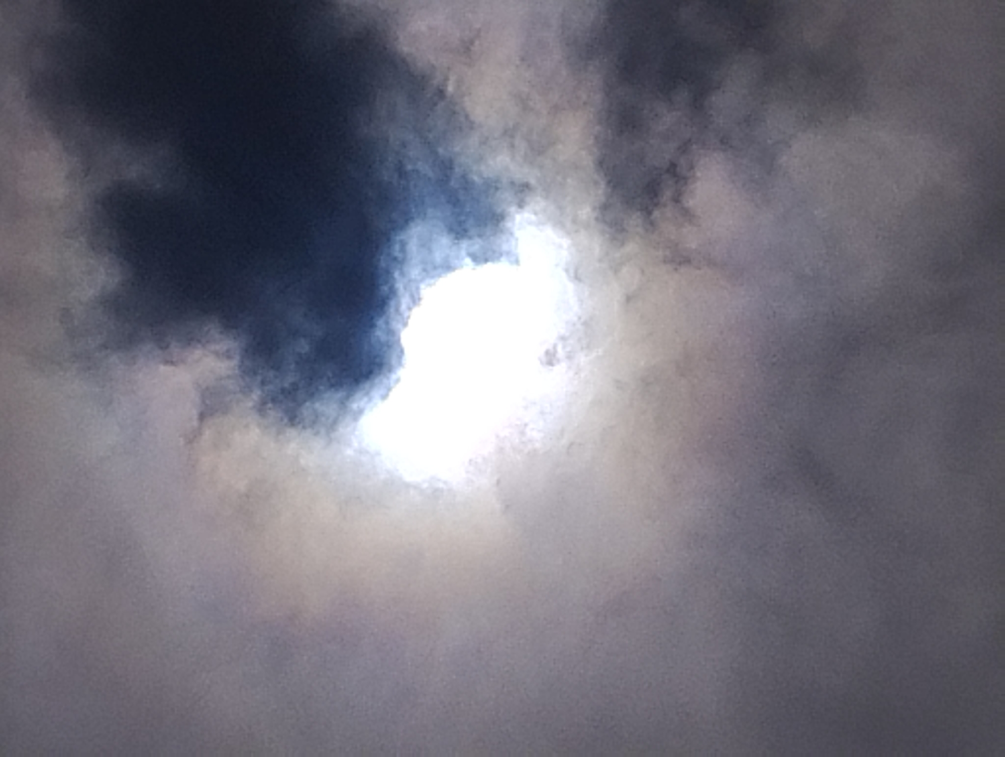 The image is a photograph of a sky with clouds. The sun is visible behind the clouds, creating a bright, luminous area in the center of the frame. Darker clouds are present on the right side of the image, contrasting with the lighter clouds and the bright sun. The overall impression is of a cloudy sky with the sun attempting to break through. The photograph appears to be taken from a low angle, looking upwards.