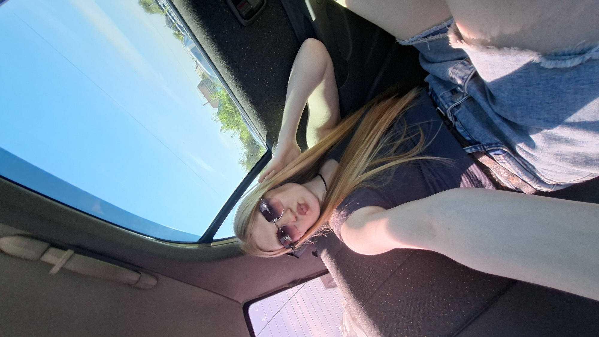 A low-angle shot taken from inside a vehicle. A young, light-skinned woman with long blonde hair and sunglasses is the primary subject. She is wearing a dark t-shirt and ripped denim shorts. She is lying on the car seat. The car has a sunroof, through which a bright blue sky and building is visible. The car's interior is dark, with a textured black fabric. The setting appears to be Прикамский, Russia, but this is inferred from the prompt, not explicitly visible in the image.
