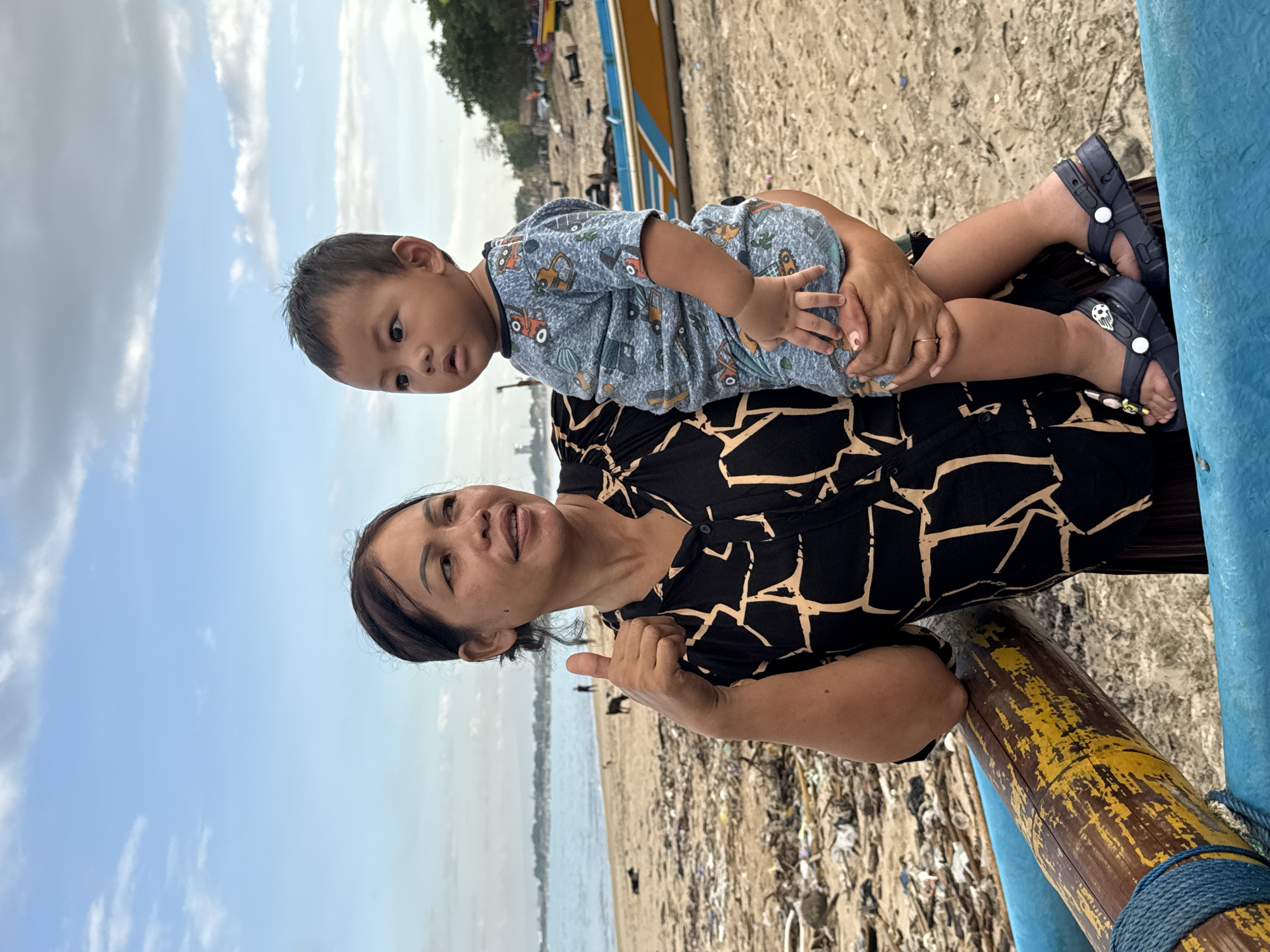 A woman and a child are on a beach near Kuta Selatan, Indonesia. The woman is holding the child, who is wearing a blue outfit with a car pattern. The woman has dark hair and is wearing a black and white patterned shirt. She is giving a thumbs-up. The beach is littered with trash. Behind them are boats and trees. The sky is blue with clouds.