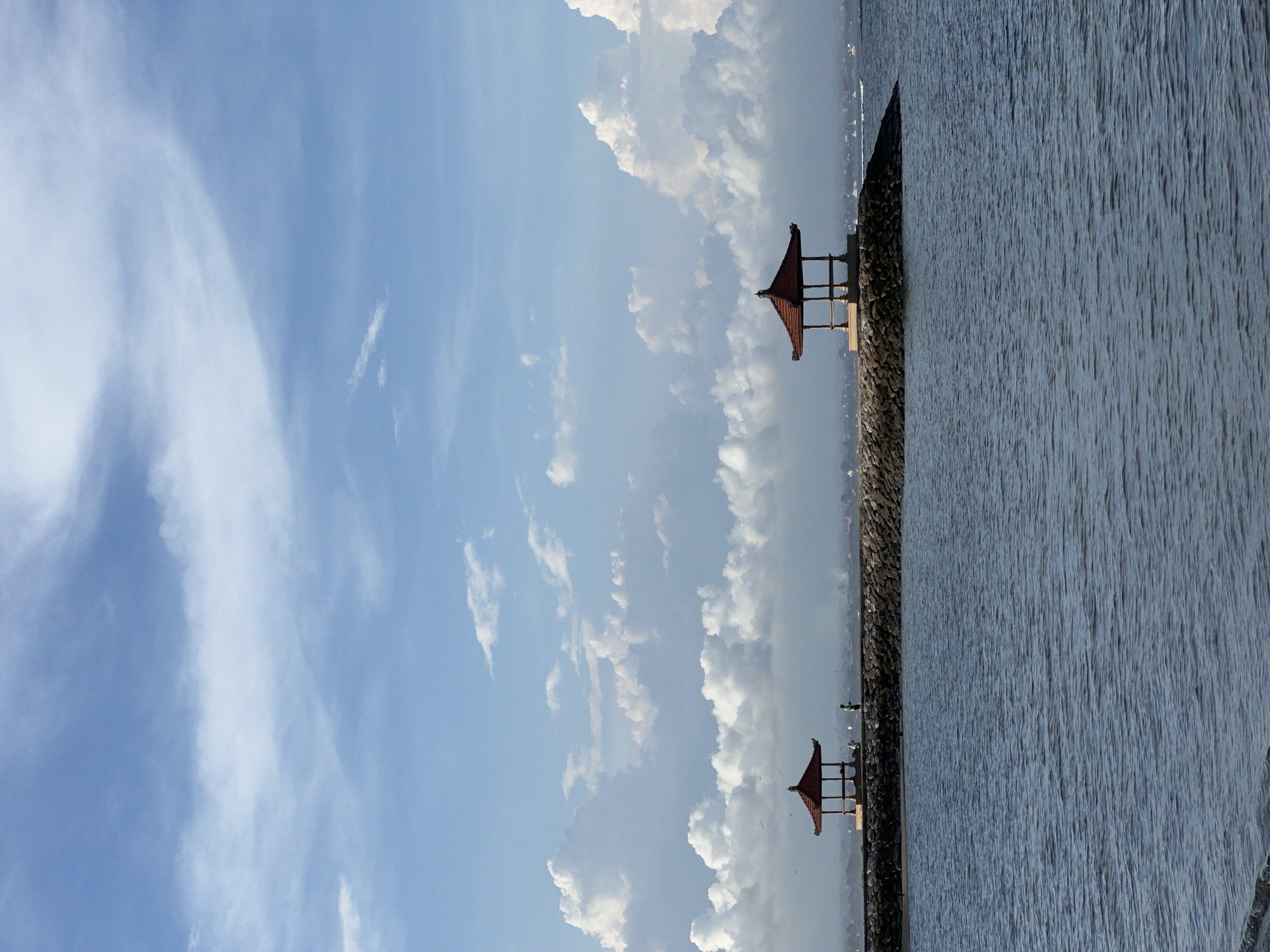 The image captures a scene at Matahari Terbit Beach in Denpasar, Indonesia. The view is dominated by a clear sky with puffy clouds, taking up the left half of the frame. Adjacent to the sky, on the right, is a body of water, its surface reflecting the light and displaying a slightly textured appearance. A dark, rocky breakwater separates the sky and clouds from the water. Two small, traditional Balinese pavilions with red roofs are situated atop the breakwater.