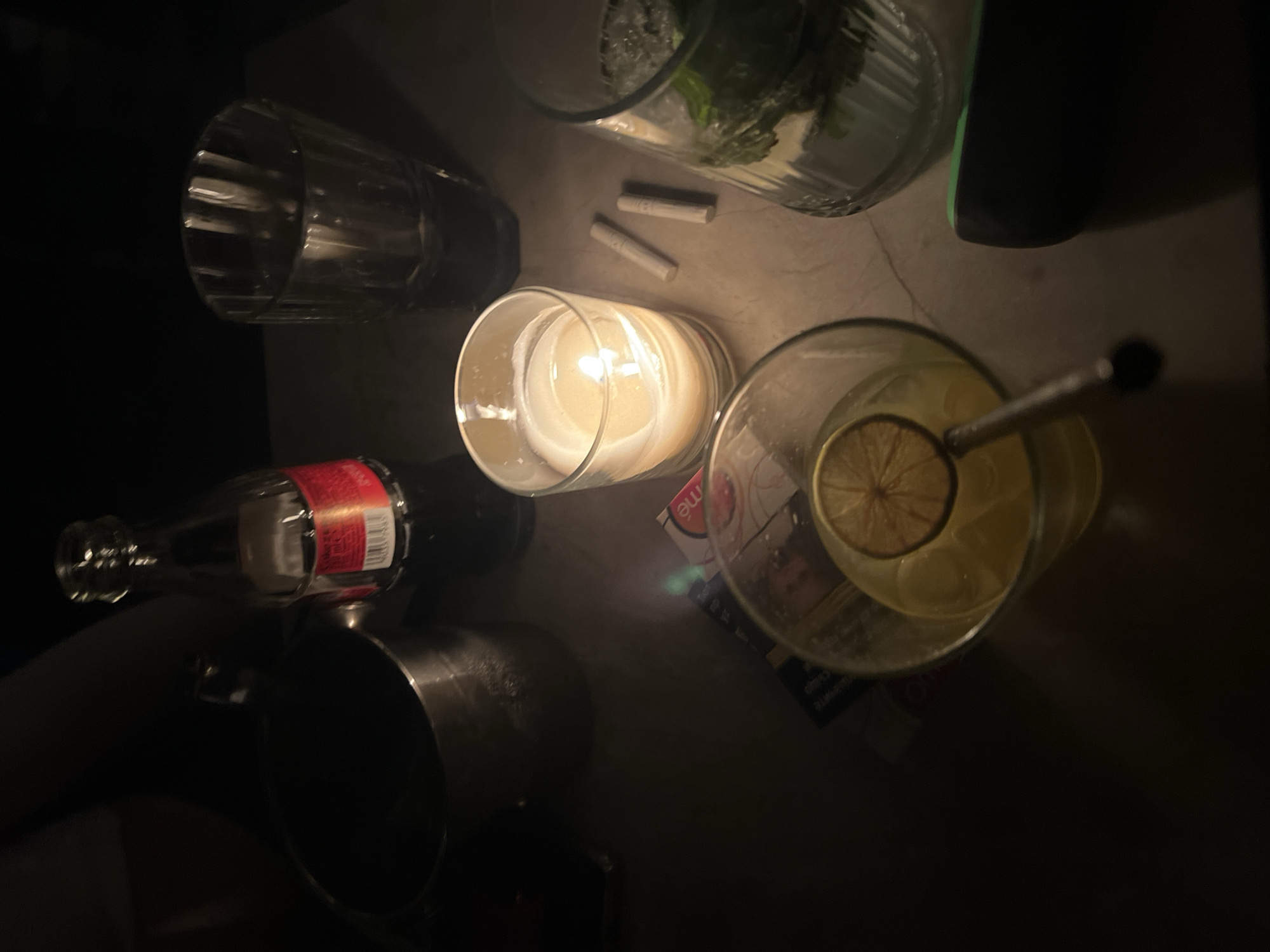 The image shows a dimly lit tabletop scene in an indoor environment. The following items are arranged on the surface: a cylindrical candle in a glass container with a lit flame, two unlit cigarettes, a bottle of Coca-Cola lying on its side, an empty glass with a ribbed pattern, a stainless steel mug, a tall glass containing ice and what appears to be mint leaves, and a glass filled with a beverage and a slice of lemon with a straw. There is also a small rectangular object that could be a card. Due to the limited information and low lighting, a precise location in Plovdiv, Bulgaria cannot be determined.