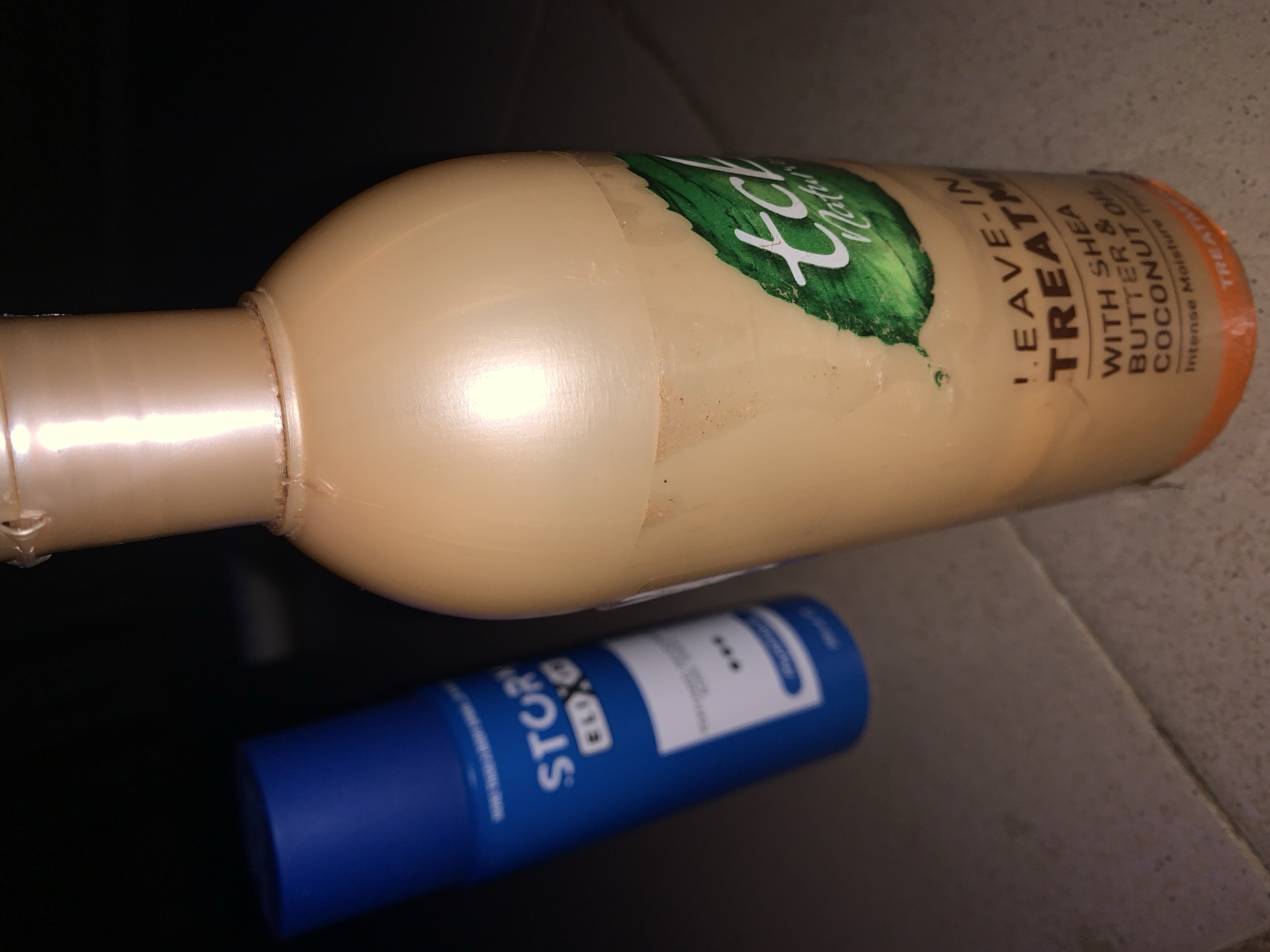 The image shows two bottles on a tiled surface. In the foreground, a pale orange bottle of "tcb Naturals LEAVE-IN TREATMENT WITH SHEA BUTTER & COCONUT OIL" is visible. The label also indicates "Intense Moisture Treatment." To the left and slightly behind, a blue bottle with a white label and the text "STORY ELIXO" is present. The lighting suggests an indoor setting, possibly a bathroom or a retail shelf, with no other people or distinguishing environmental features visible.