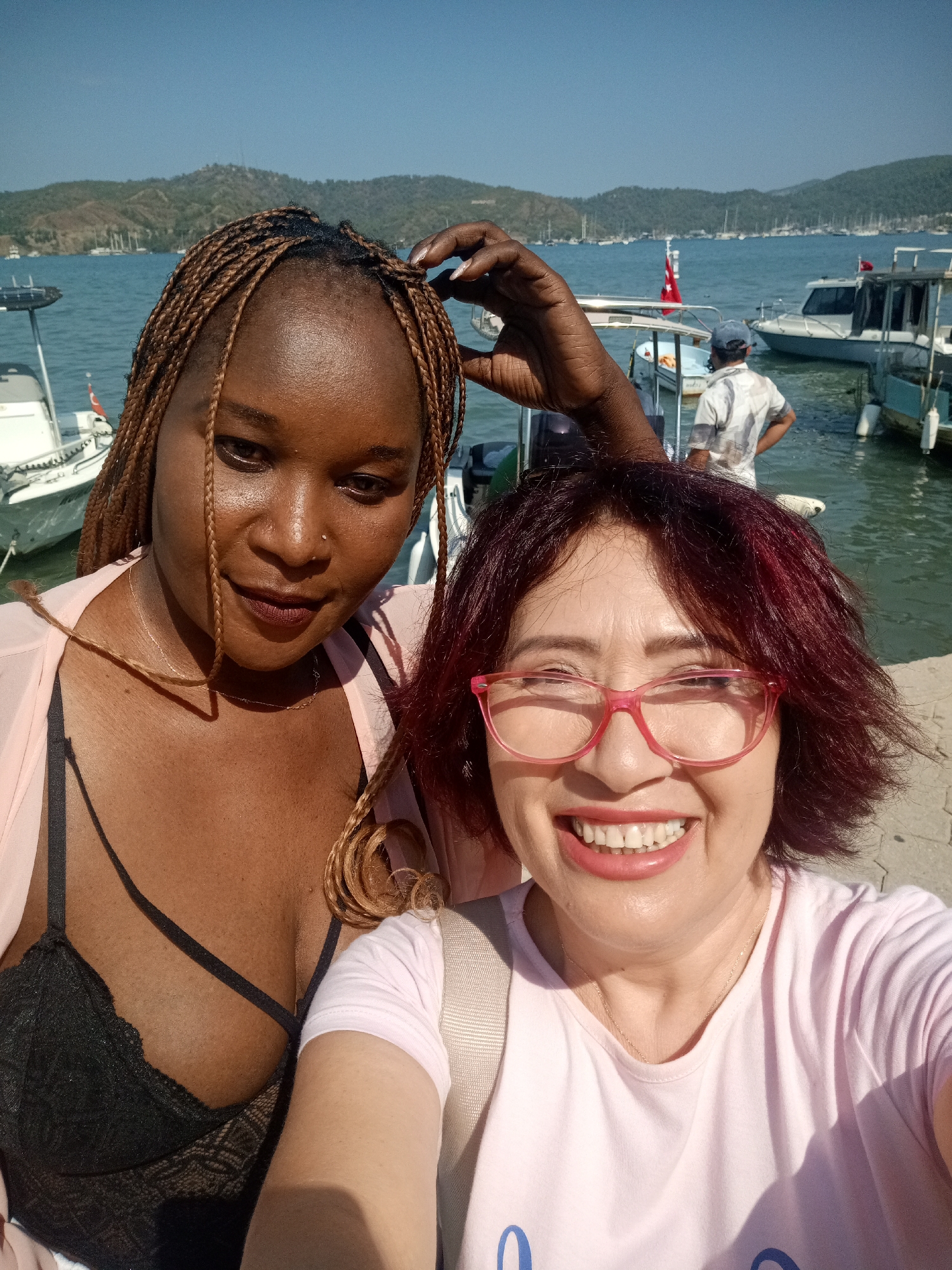Two women are taking a selfie on a sunny day by the water. The woman on the left has dark skin and braided hair. She is wearing a black bra and a sheer pink cover-up. The woman on the right has fair skin and short, dark red hair. She is wearing glasses with pink frames and a light pink t-shirt. Both women are smiling. In the background, there are boats docked in the water, and hills covered in trees rise behind them. A man in a patterned shirt and a blue cap is visible standing on one of the boats. The scene appears to be a harbor or marina in Fethiye, Turkey, indicated by the Turkish flags on the boats. The water is a clear blue, and the sky is bright and cloudless. The ground where the women are standing appears to be paved.