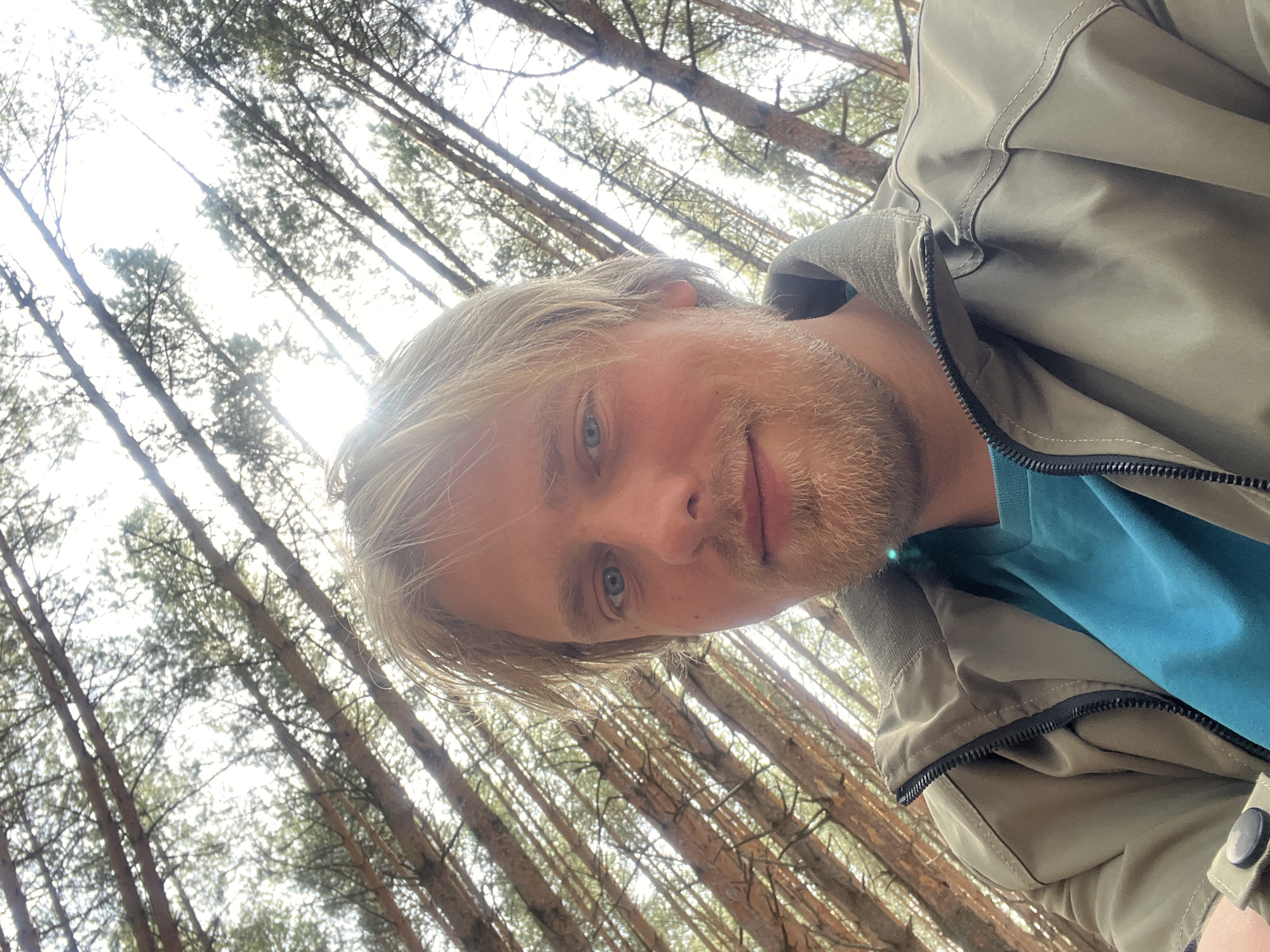 The image is a selfie of a man with blond hair and a beard. He is wearing a green jacket over a blue shirt. The background consists of tall pine trees, suggesting he is in a forest. The location is possibly in or near Chaikovsky, Russia, given the prompt. The trees are dense, with the sky visible through the canopy. The camera is pointed upwards, capturing the trees and the sky behind the man.