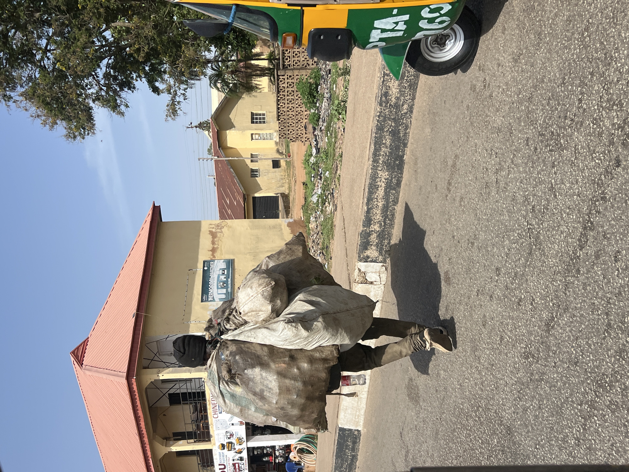 A man is walking down a paved street in Jos, Nigeria. He carries a large load of bundled, dirty, and heavy-looking sacks on his back. The sacks appear to be quite cumbersome, obstructing a clear view of the man himself. His attire includes shoes and what may be dark-colored pants. In the background are low-rise buildings, including one with a reddish-orange roof. A motorized tricycle (keke) is partially visible in the upper right corner. Debris is scattered between buildings along the roadside. The sky is clear and bright, indicating a sunny day.