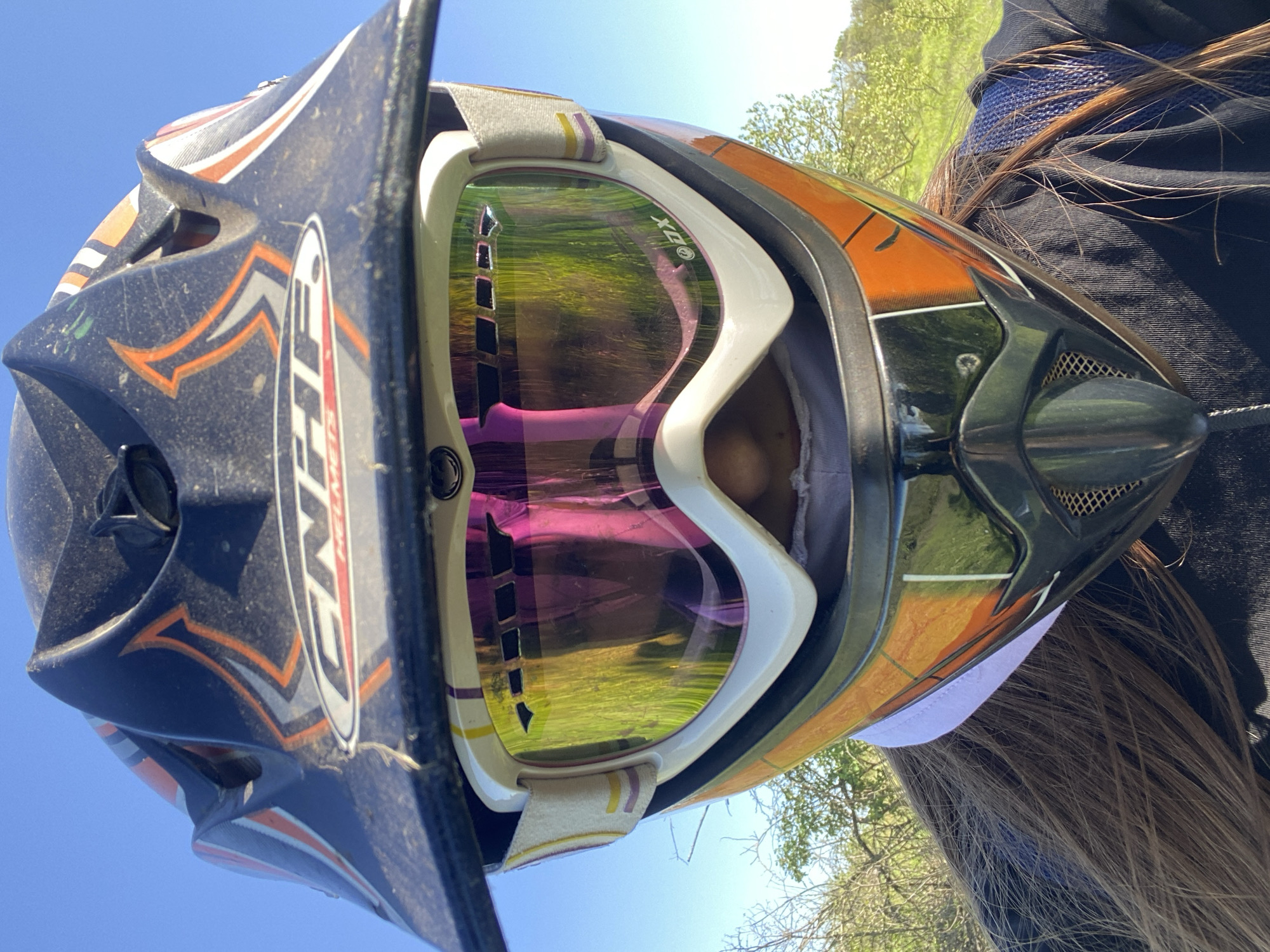 A close-up shot looking upwards at a person wearing a motocross helmet with goggles. The helmet is predominantly black, orange, and white, with racing style graphics. The goggles have a white frame and a reflective lens that appears to reflect a grassy landscape with trees. The sky is visible as a bright blue backdrop. Part of the person's brown hair and dark clothing is visible on the right side of the frame. The location is Konstantinovo, Bulgaria.